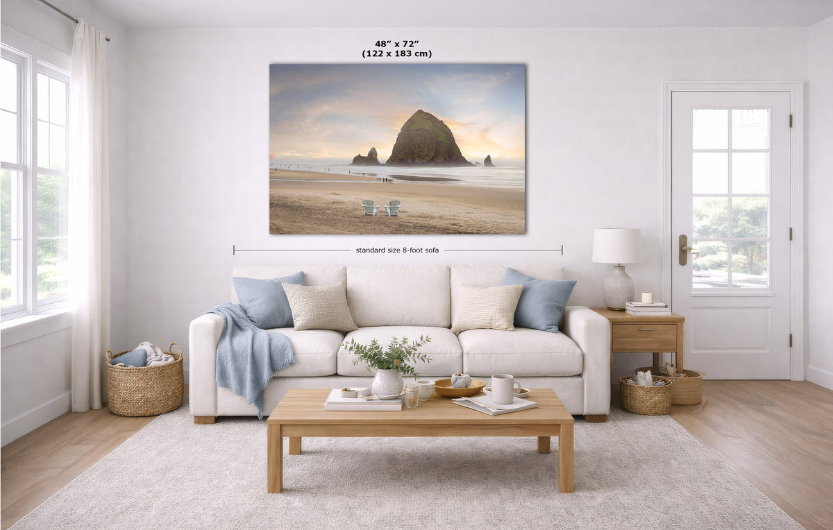 A large horizontal canvas print of Haystack Rock on Cannon Beach in Oregon displayed on a white wall in a modern, bright living room. The image includes dimension lines and text indicating the size is 48" x 72" (122 x 183 cm) with a standard 9-foot height.