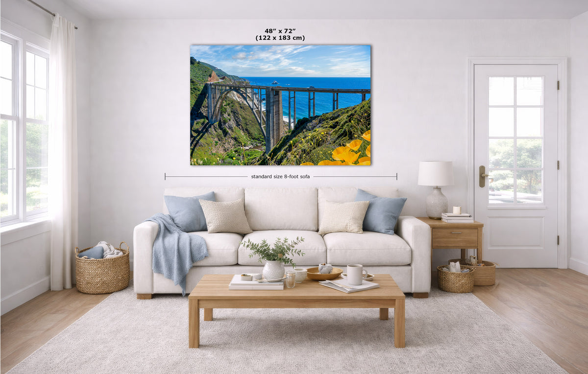 A wall art print showing a scenic bridge over blue water and green hills, displayed above a white sofa in a bright modern living room. Measurement labels indicate the print size as 48 x 72 inches.