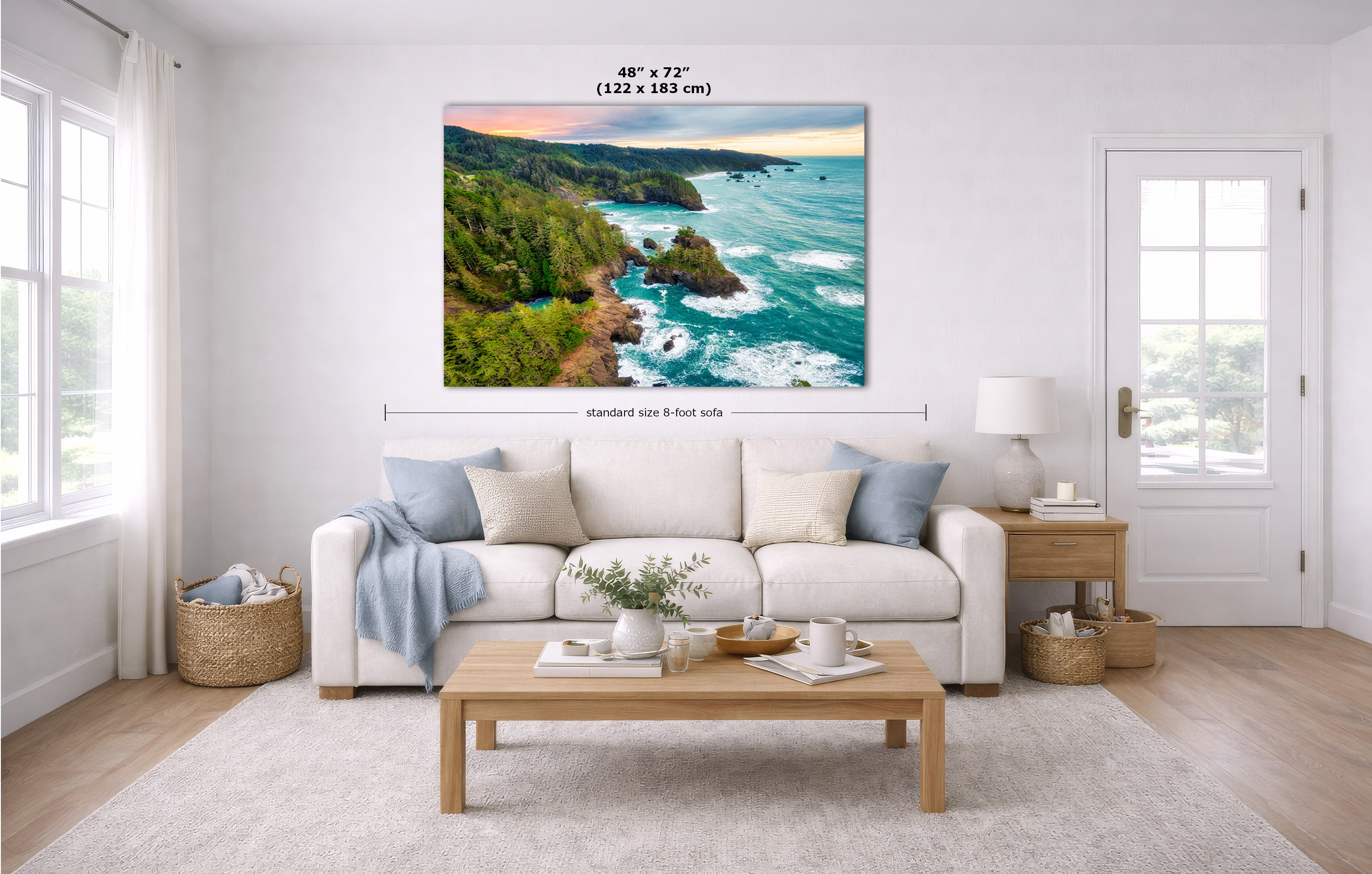 A large rectangular coastal landscape wall art print hangs above a white sofa in a bright living room. The artwork shows turquoise ocean water, rocky coastline, green cliffs, and a sunset sky.