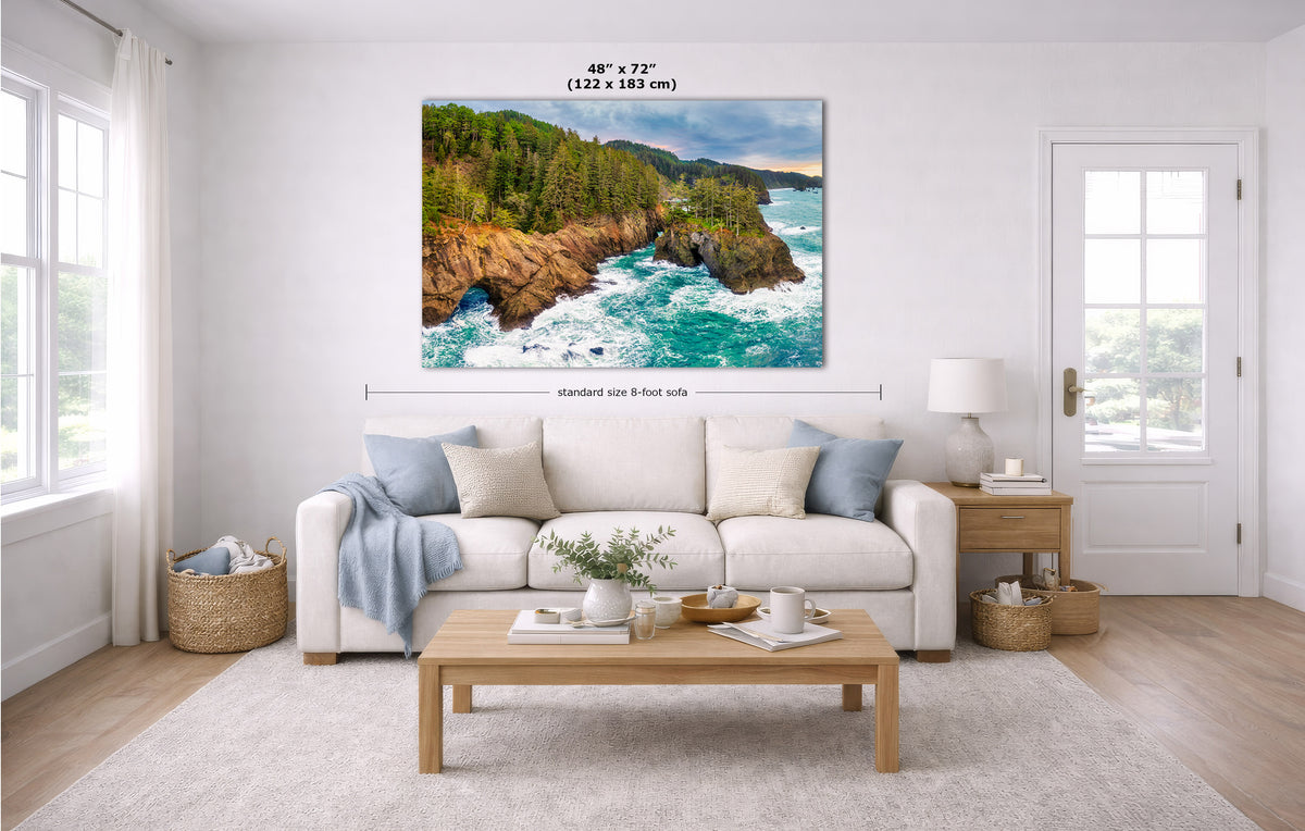 A large square wall art print showing a coastal rocky shoreline with turquoise water and green trees, displayed above a white sofa in a bright living room. Measurement lines and text indicate the print size as 48 x 72 inches.