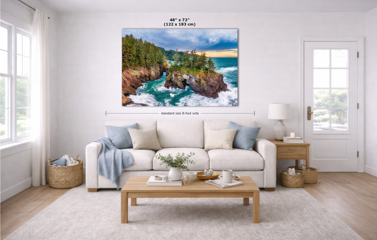 A large coastal landscape wall art print hangs above a white sofa in a bright living room. The artwork shows rocky cliffs, turquoise water, green trees, and a sunset sky.