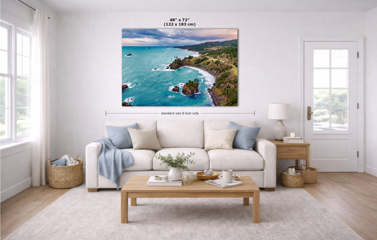 A large coastal landscape wall art print hangs above a white sofa in a bright living room. The artwork shows turquoise ocean water, rocky coastline, green cliffs, and a sunset sky.