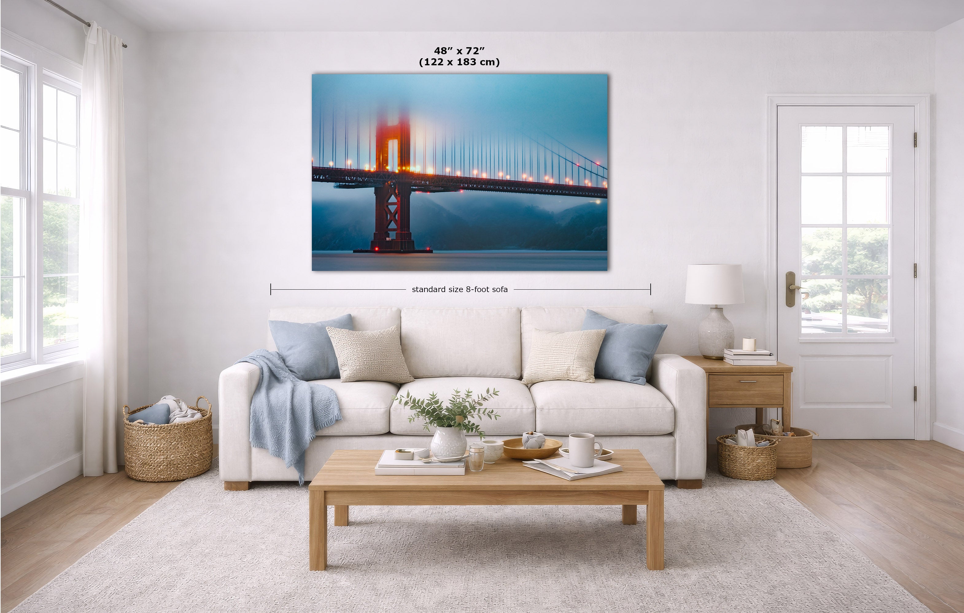 A wall art print of the Golden Gate Bridge hanging above a white sofa in a bright, modern living room. The print shows the bridge at dusk with blue water and sky, orange-red bridge structure, and a warm glowing light.