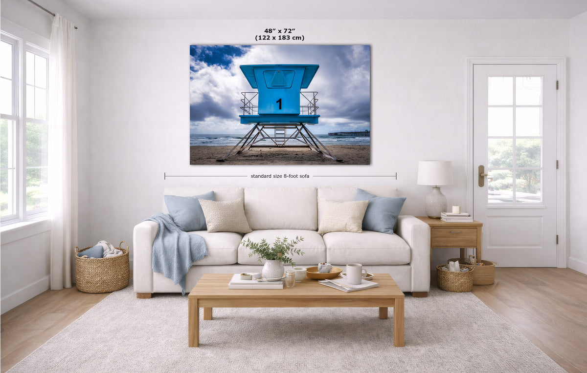 A large wall art print showing a blue lifeguard tower on a beach under a cloudy sky, displayed above a white sofa in a bright modern living room. Measurement labels for the print size are overlaid on the image.