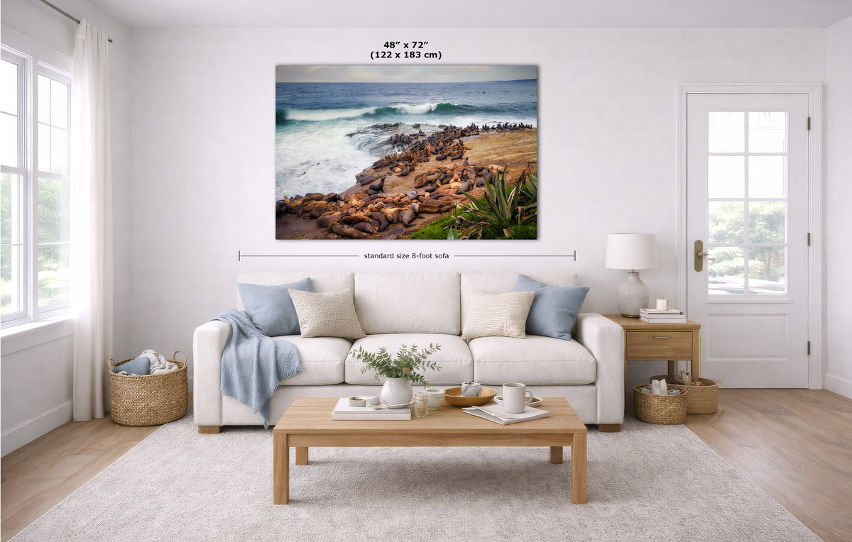 A large coastal landscape wall art print hangs above a white sofa in a bright modern living room. The artwork shows a rocky beach with ocean waves and greenery.