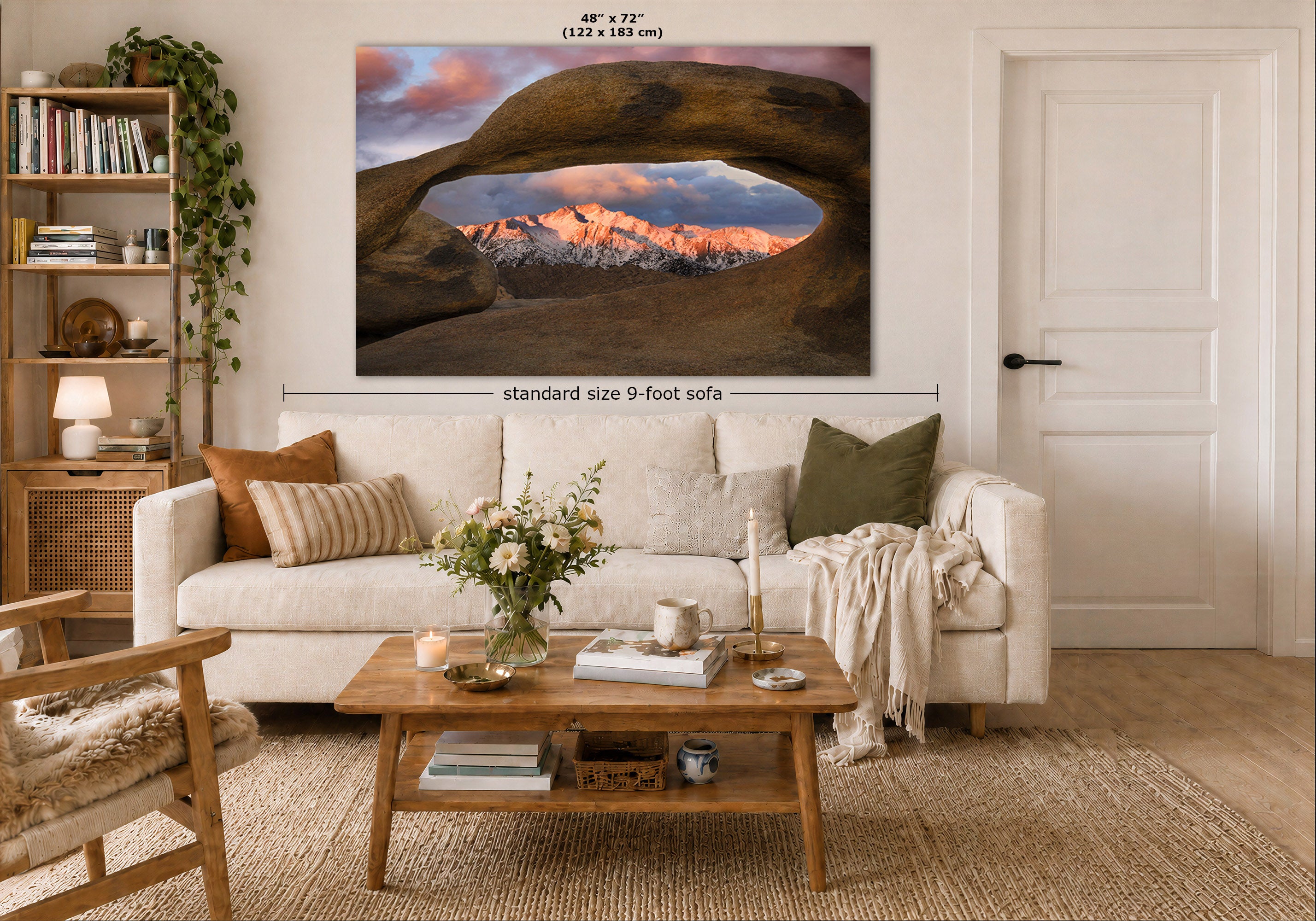 A large wall art print showing a desert rock arch over a mountain range with pink clouds, displayed above a white sofa in a styled living room. Measurement text indicates the print is 48 x 72 inches and the sofa is standard size 9-foot.