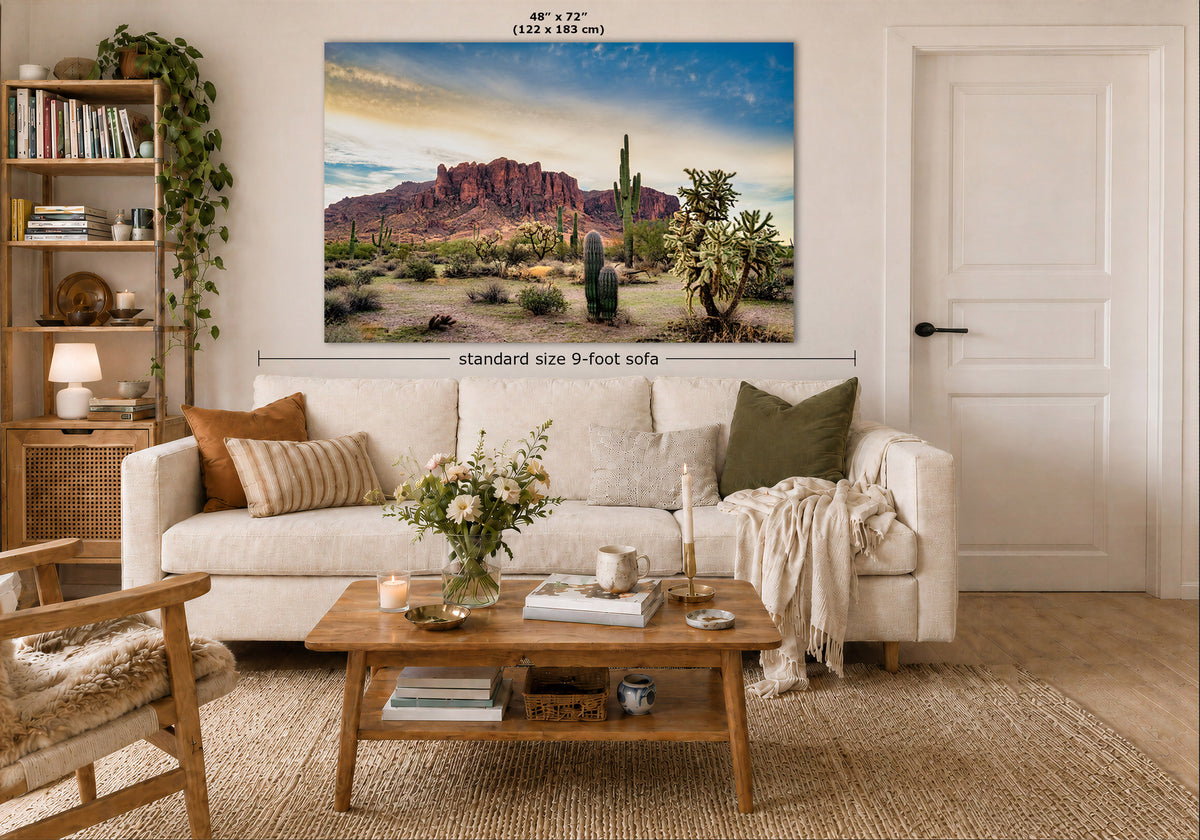 A large rectangular wall art print showing a desert landscape with red rock formations, cacti, and a blue sky, displayed above a white sofa in a styled living room. Measurement text and arrows indicate the print size as 48 x 72 inches (122 x 183 cm) and notes 'standard size 9-foot sofa'.