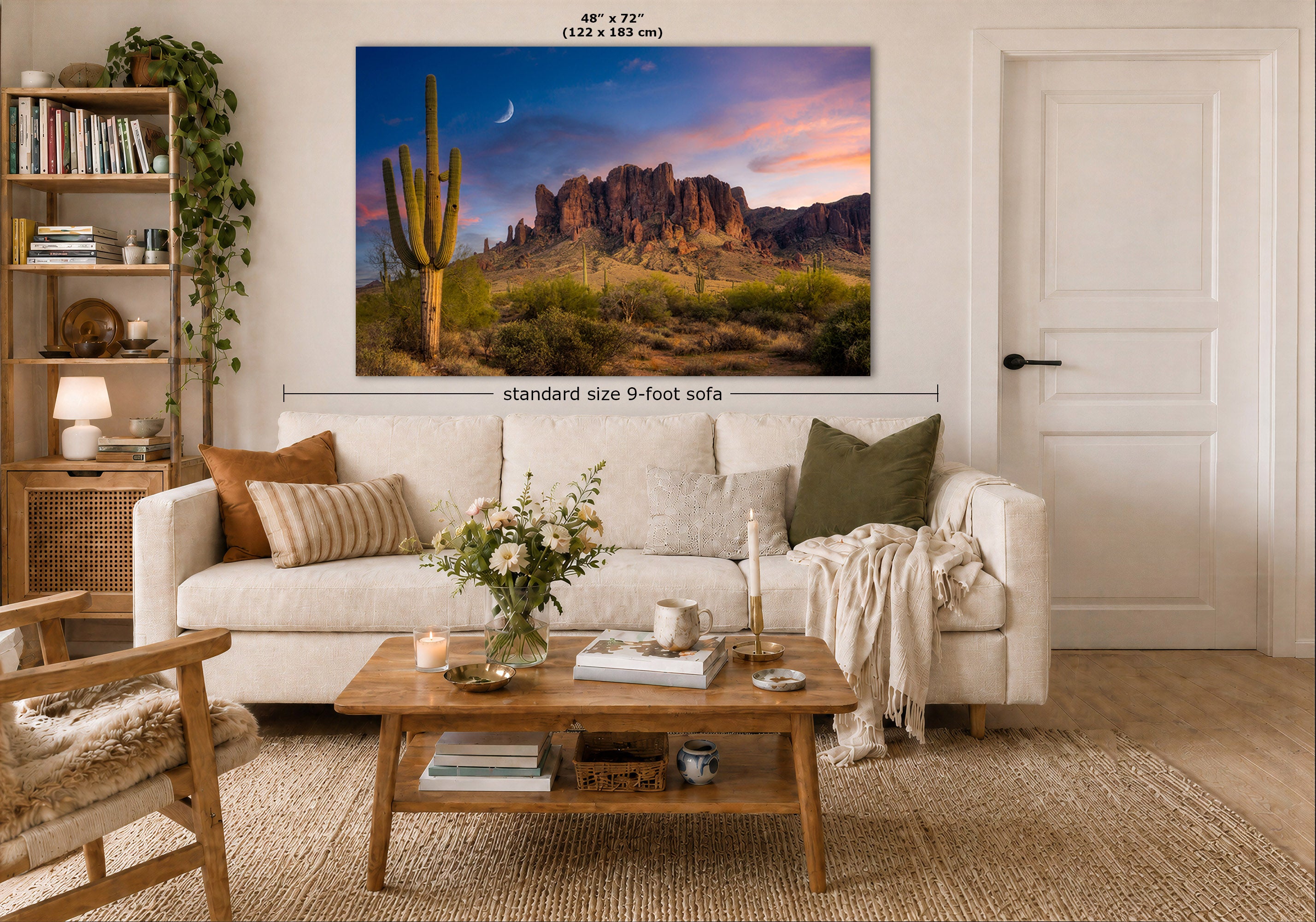 A large desert landscape wall art print hangs above a white sofa in a styled living room. The artwork shows a cactus, rocky mountains, and a sunset sky.