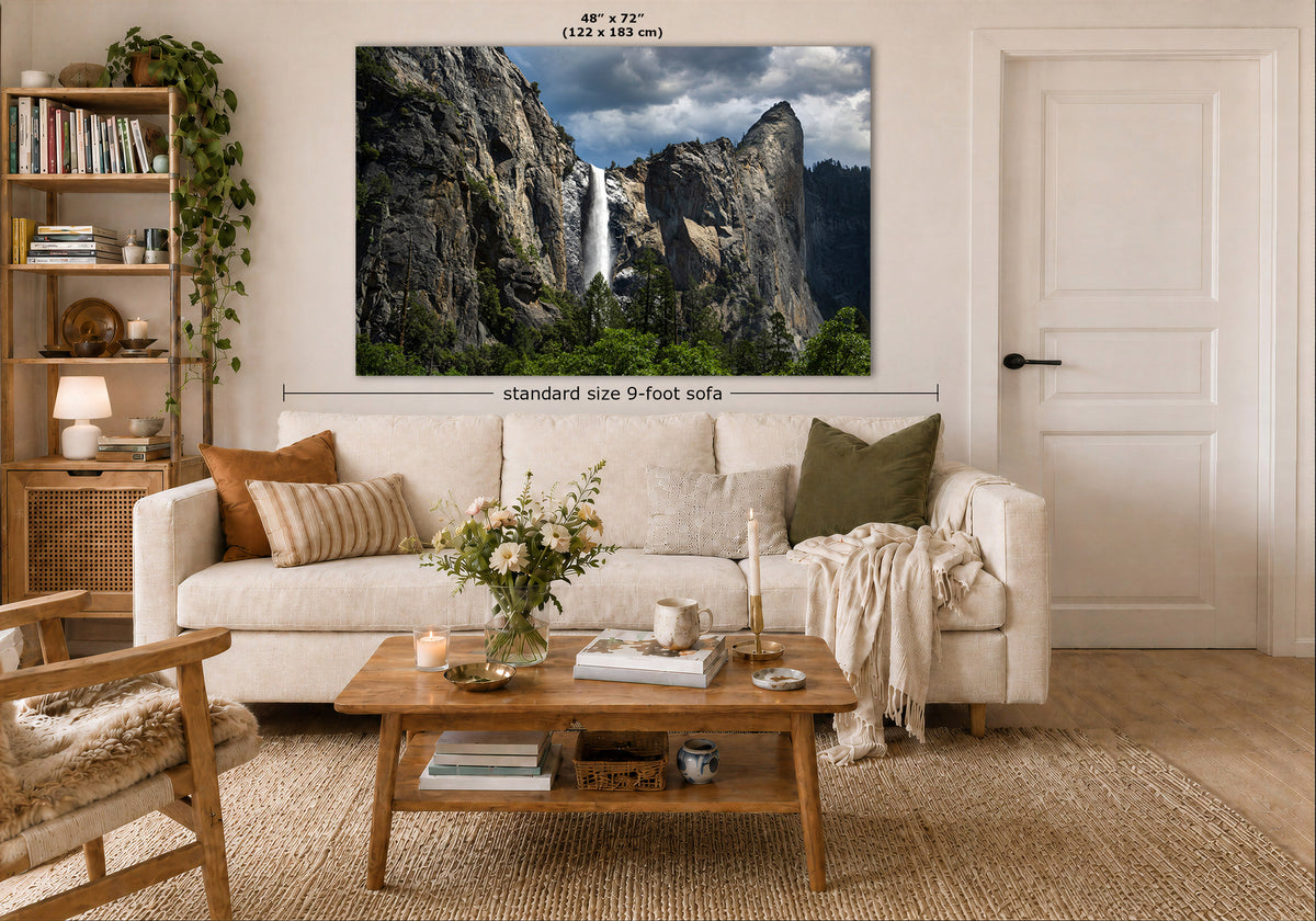 A large framed wall art print showing a waterfall and rocky mountain landscape is displayed above a cream sofa in a styled living room. White dimension lines and text indicate the print size as 48 x 72 inches, with additional room context including a coffee table, bookshelf, and door.