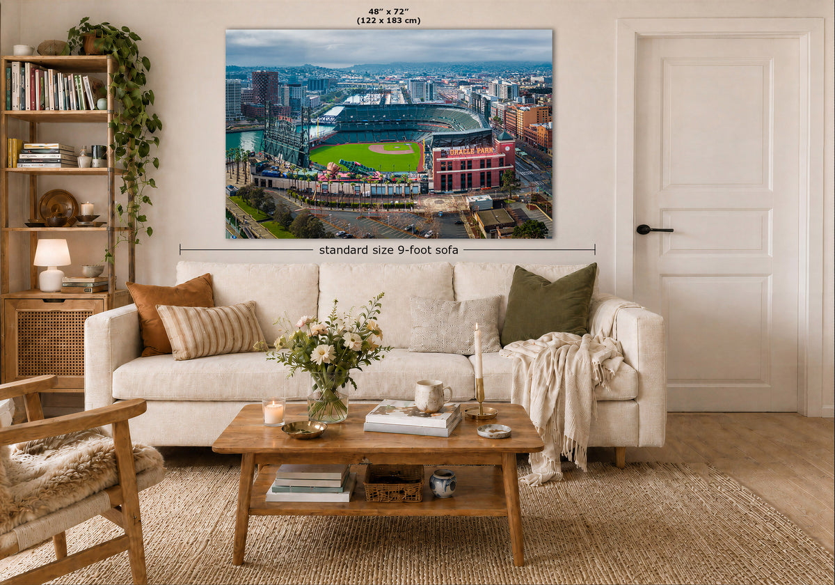 A large wall art print showing an aerial view of a city stadium and surrounding buildings, displayed above a white sofa in a styled living room. The print includes size text reading 48" x 72" and a dimension marker for a standard size 9-foot sofa.