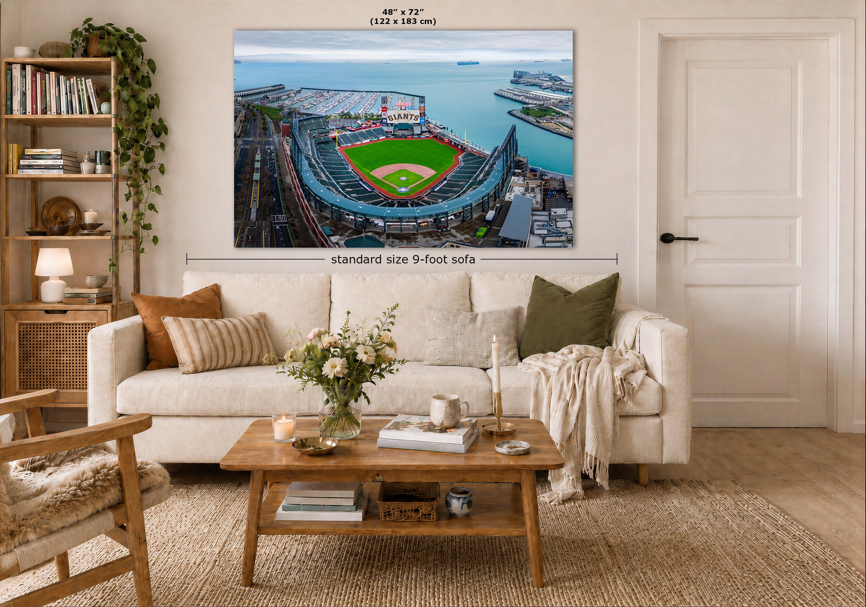 A large wall art print of a baseball stadium aerial view hangs above a white sofa in a styled living room. The print includes size markings reading 48" x 72" and standard size 9-foot sofa context, with the room showing a coffee table, pillows, bookshelf, and door.