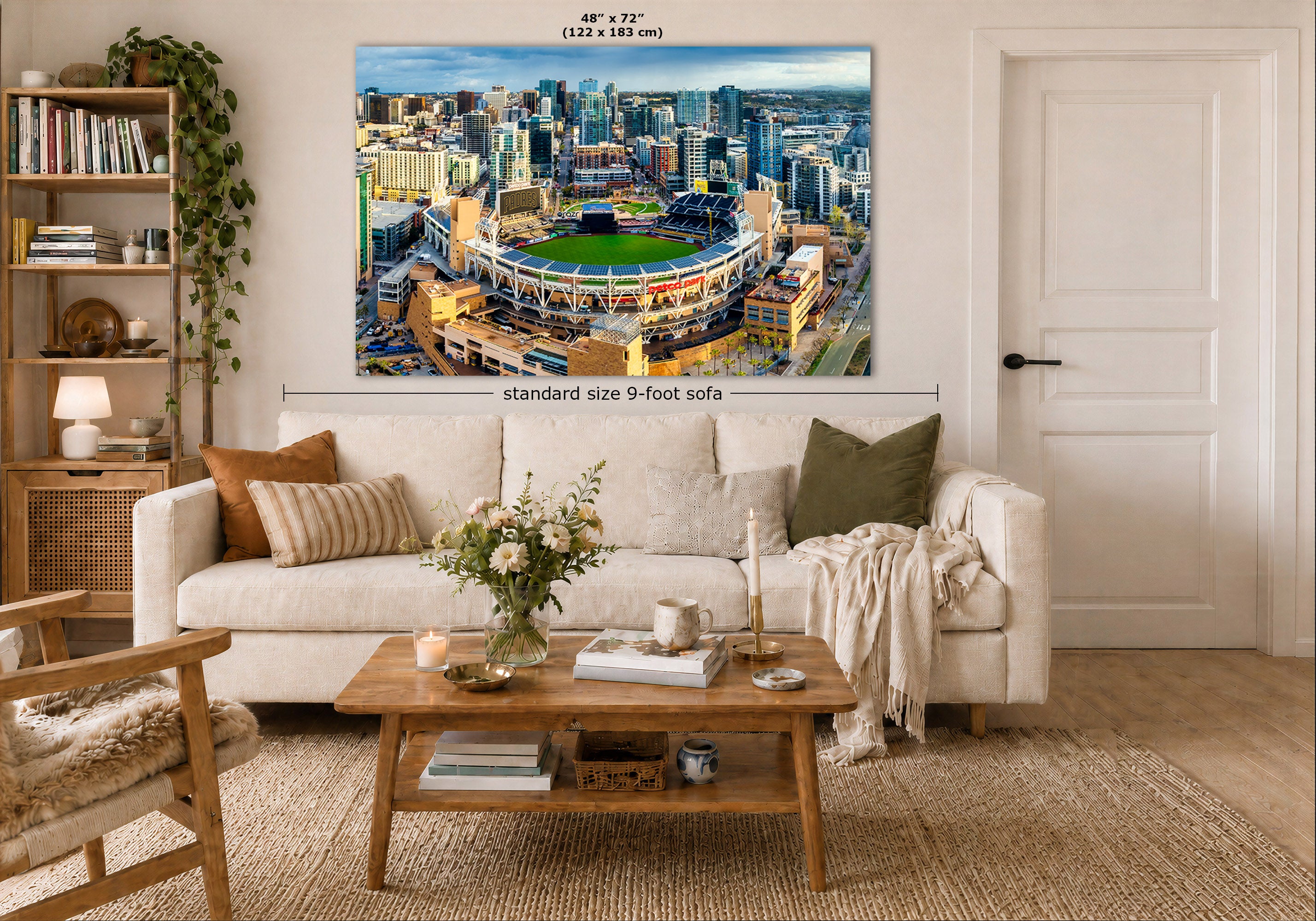 A San Diego Padres Stadium art print showing an aerial view of Petco Park baseball stadium and the surrounding San Diego city skyline, displayed above a white sofa in a styled living room. White text on the image indicates dimensions and notes that the print fits a standard 9-foot sofa.