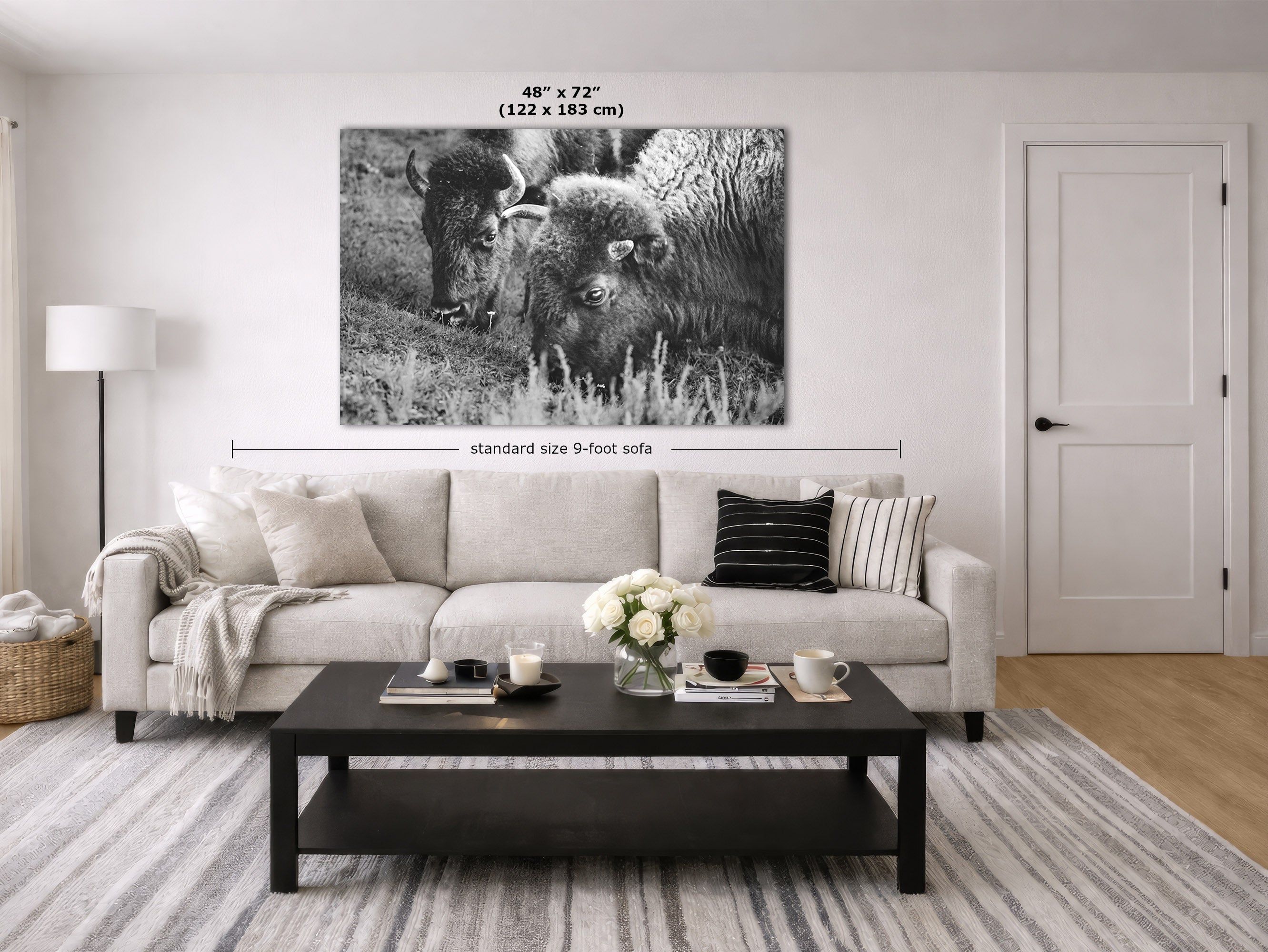 A large black and white canvas wall art print depicting two bison, shown hanging on a white wall in a modern living room mockup above a light beige sofa.