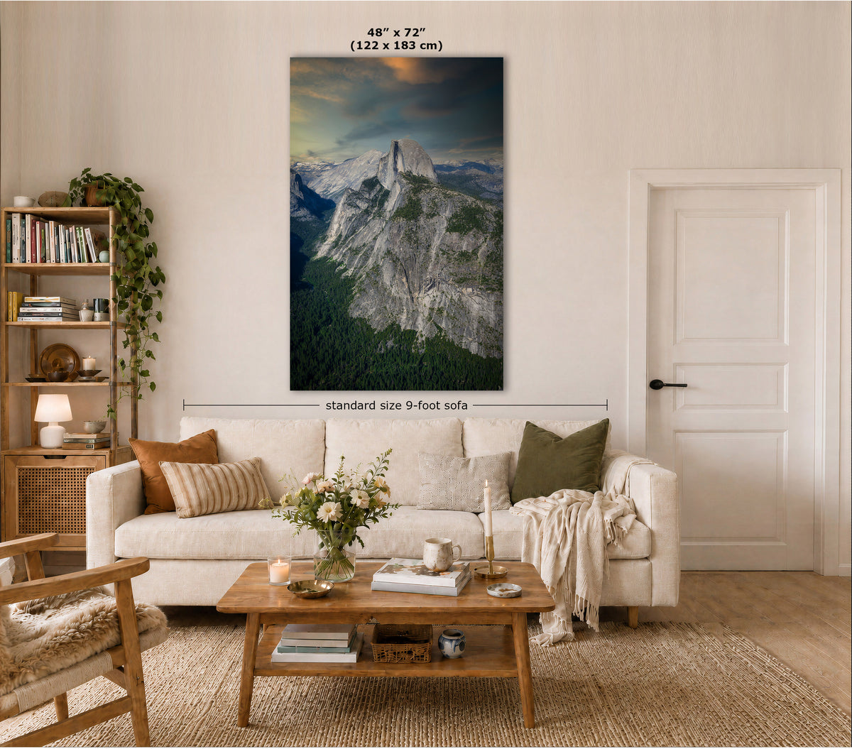 A large vertical canvas print of a mountain landscape hangs on a beige wall in a styled living room. The print shows a rocky mountain peak under a dramatic cloudy sky.