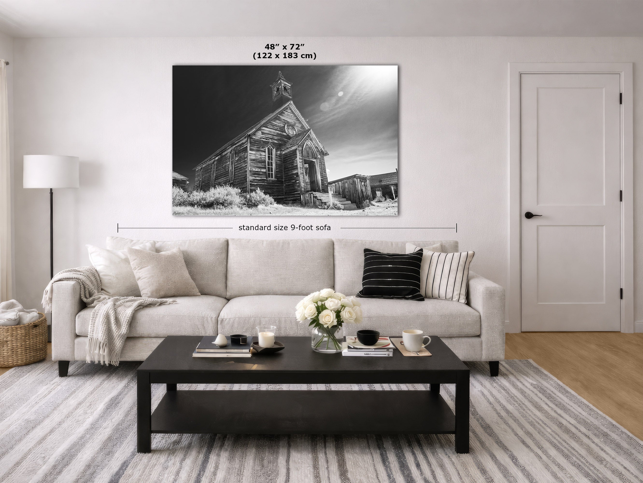 A large black and white canvas wall art print depicting an old wooden church building, shown hanging above a light beige sofa in a modern, bright living room. Text overlays indicate the print's dimensions as 48" x 72" (122 x 183 cm) and note the standard size of the sofa is 9 feet.