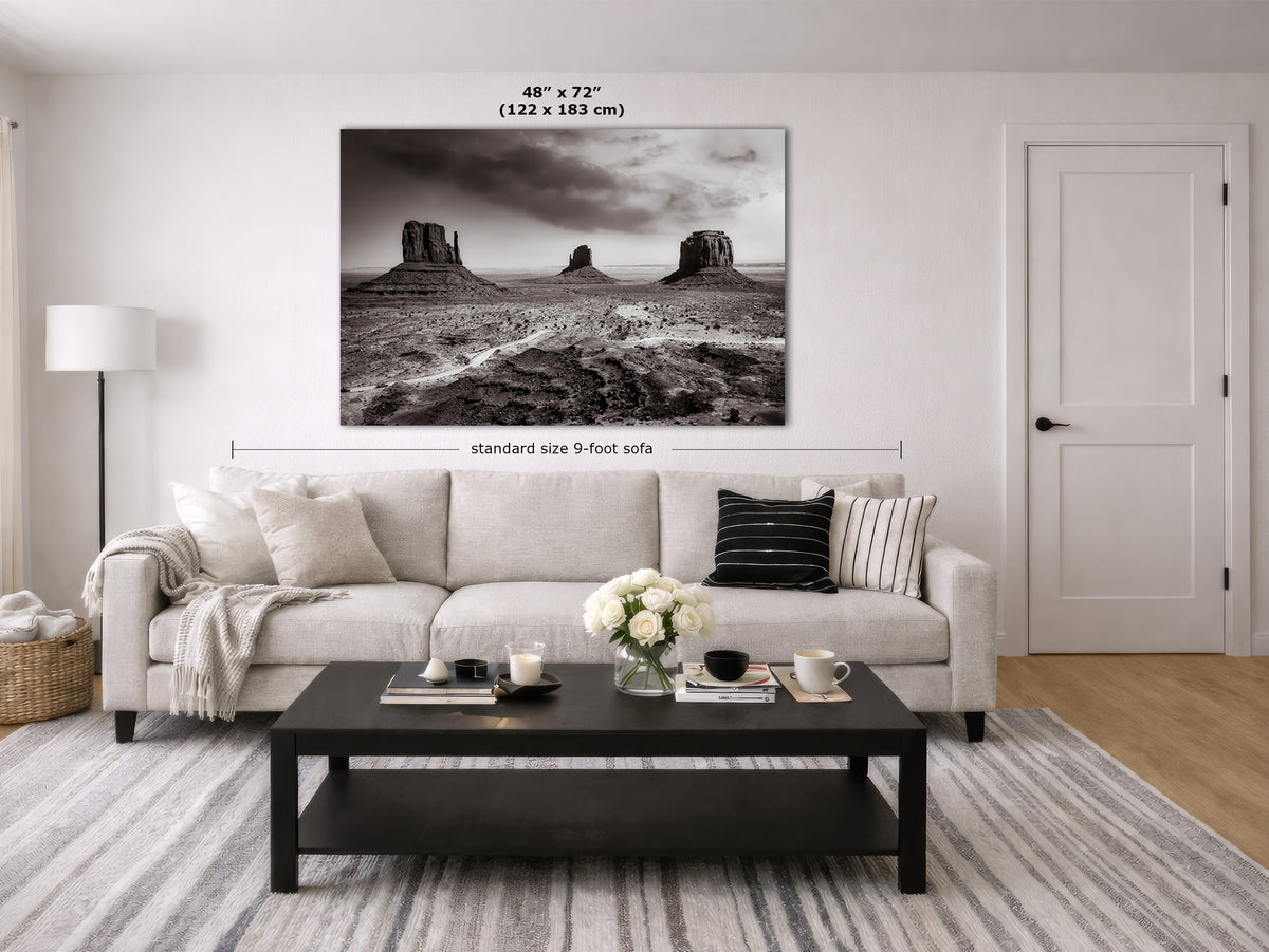 A large black and white landscape canvas print of Monument Valley is displayed on a white wall above a light beige sofa in a modern living room. The image includes dimension markers indicating the print is 48 inches by 72 inches (122 x 183 cm) and the sofa is standard size 9 feet.