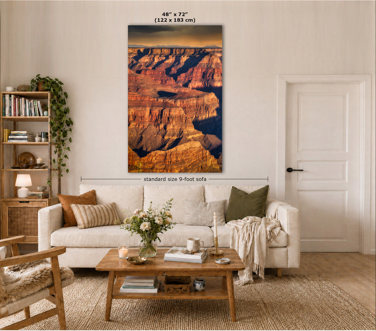 A large vertical canvas print of the Grand Canyon hangs on a beige wall in a styled living room. The print shows layered canyon cliffs in warm orange, brown, and deep blue tones.