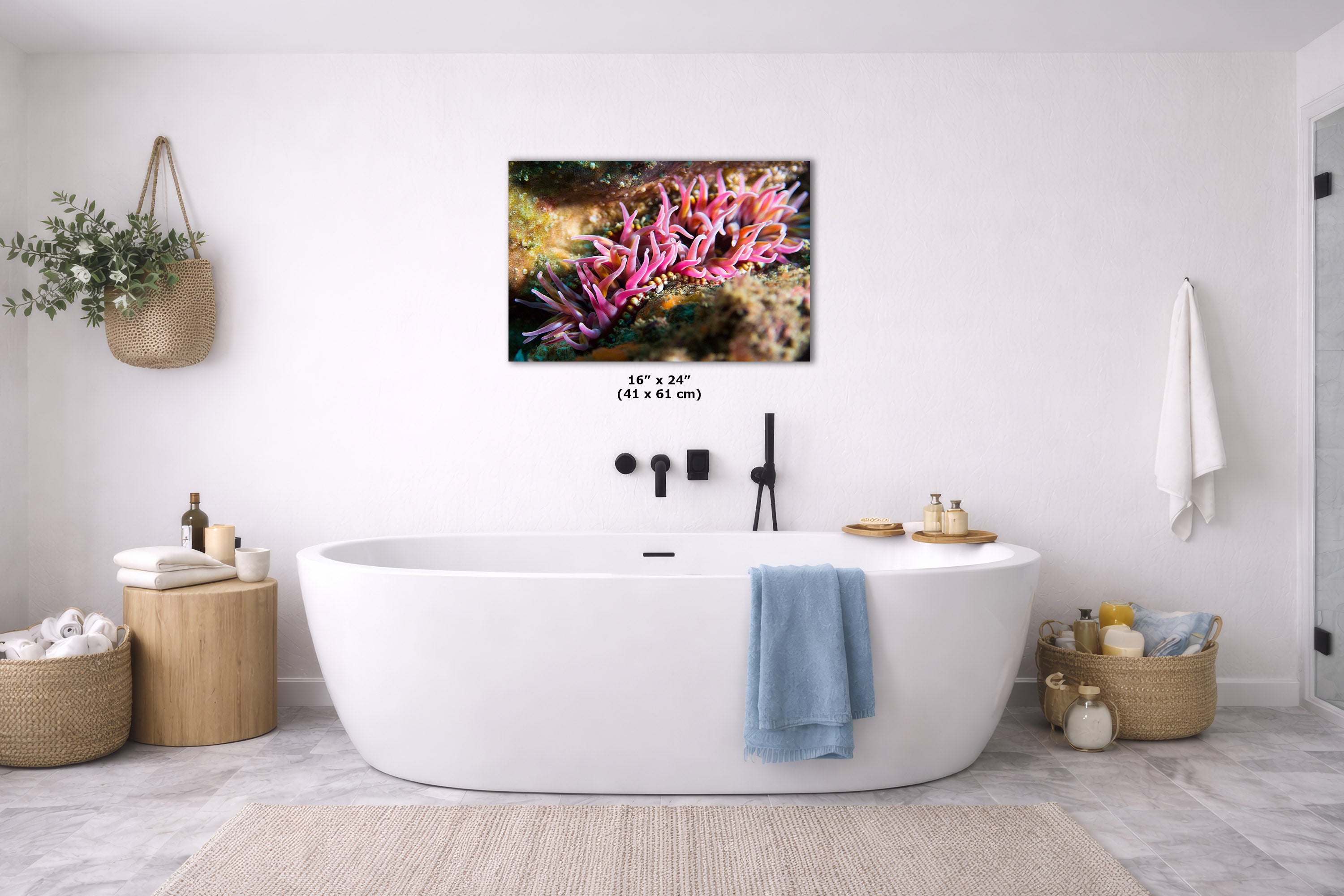 A freestanding oval bathtub in a bright white bathroom, with a colorful coral print hanging on the wall above it. The room includes a blue towel draped over the tub, woven baskets, a small side table, and a light rug on the floor.