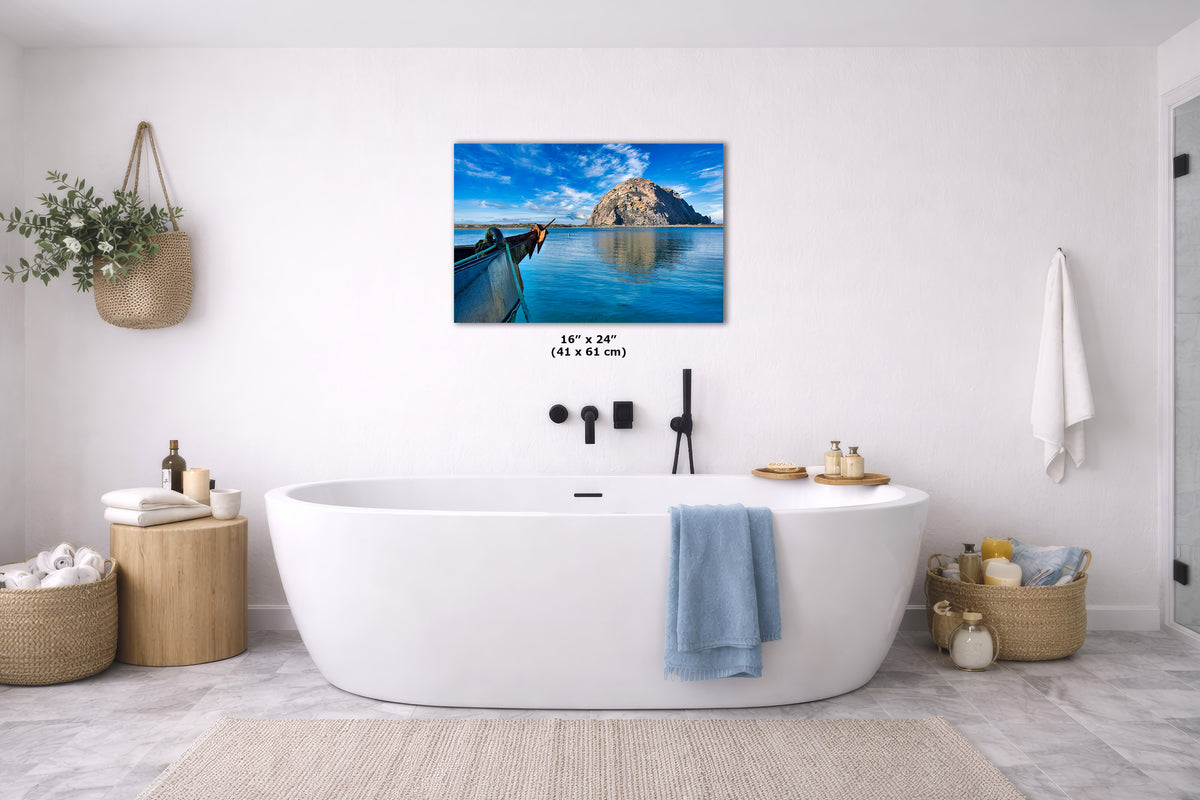 A wall art print showing an island scene with a longtail boat and blue water, displayed above a white freestanding bathtub in a bright modern bathroom. Small text below the print reads 16" x 24" (41 cm x 61 cm).