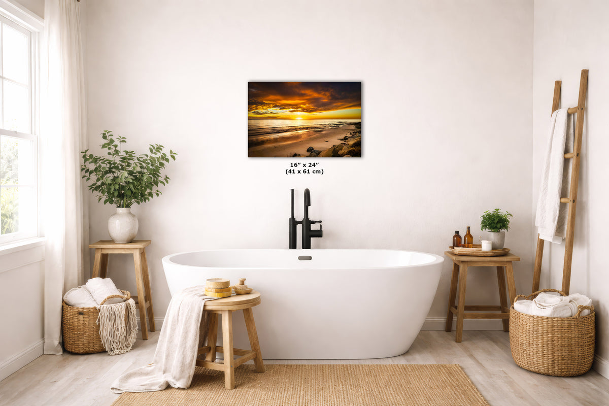 A wall art print depicting a dramatic sunset over water, styled in a bright, modern bathroom above a freestanding white bathtub. The print is shown with its dimensions printed below it.
