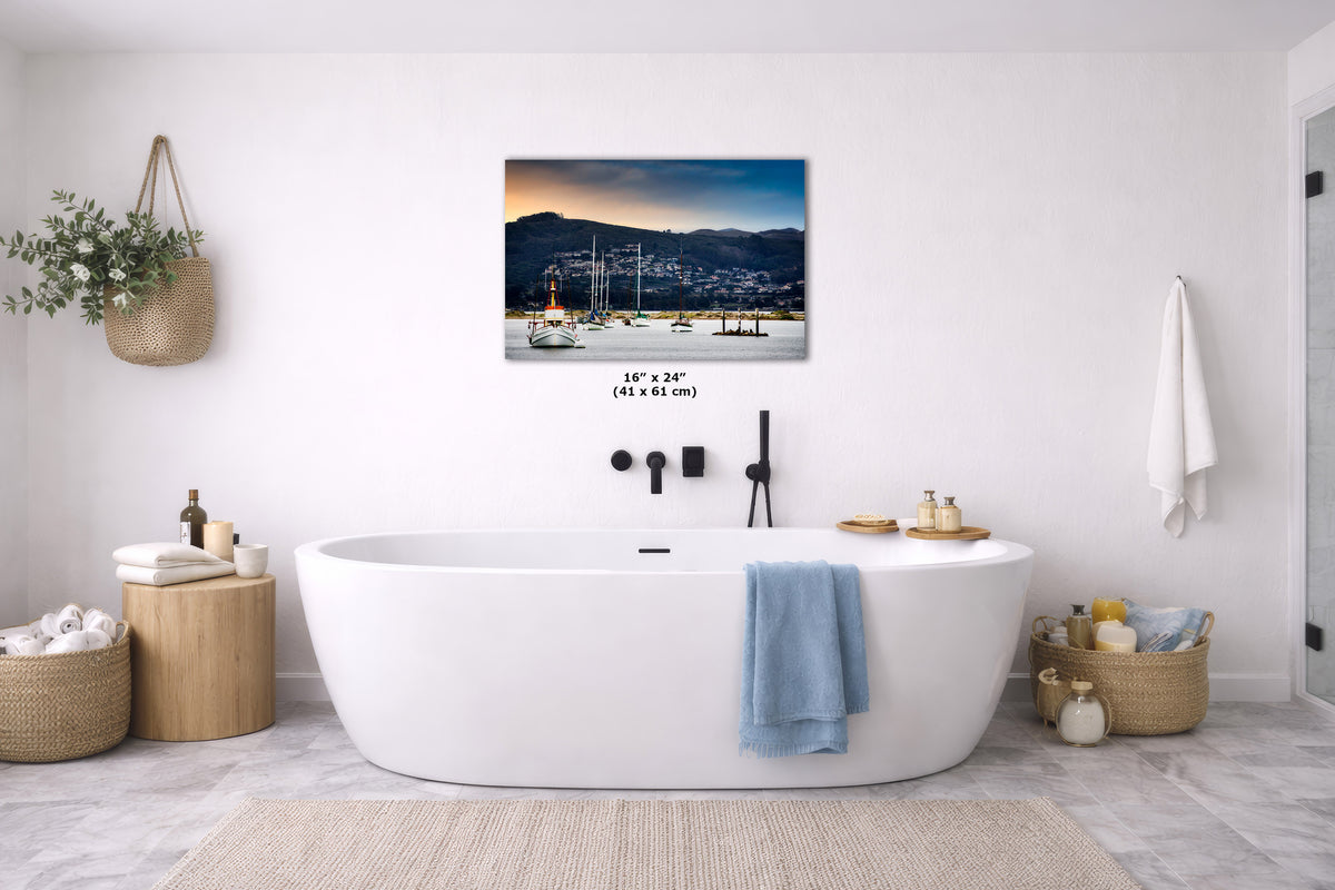 A wall art print depicting a harbor scene with boats at dusk, displayed above a white freestanding bathtub in a bright, modern bathroom. The print includes size text reading 16 x 24 in.
