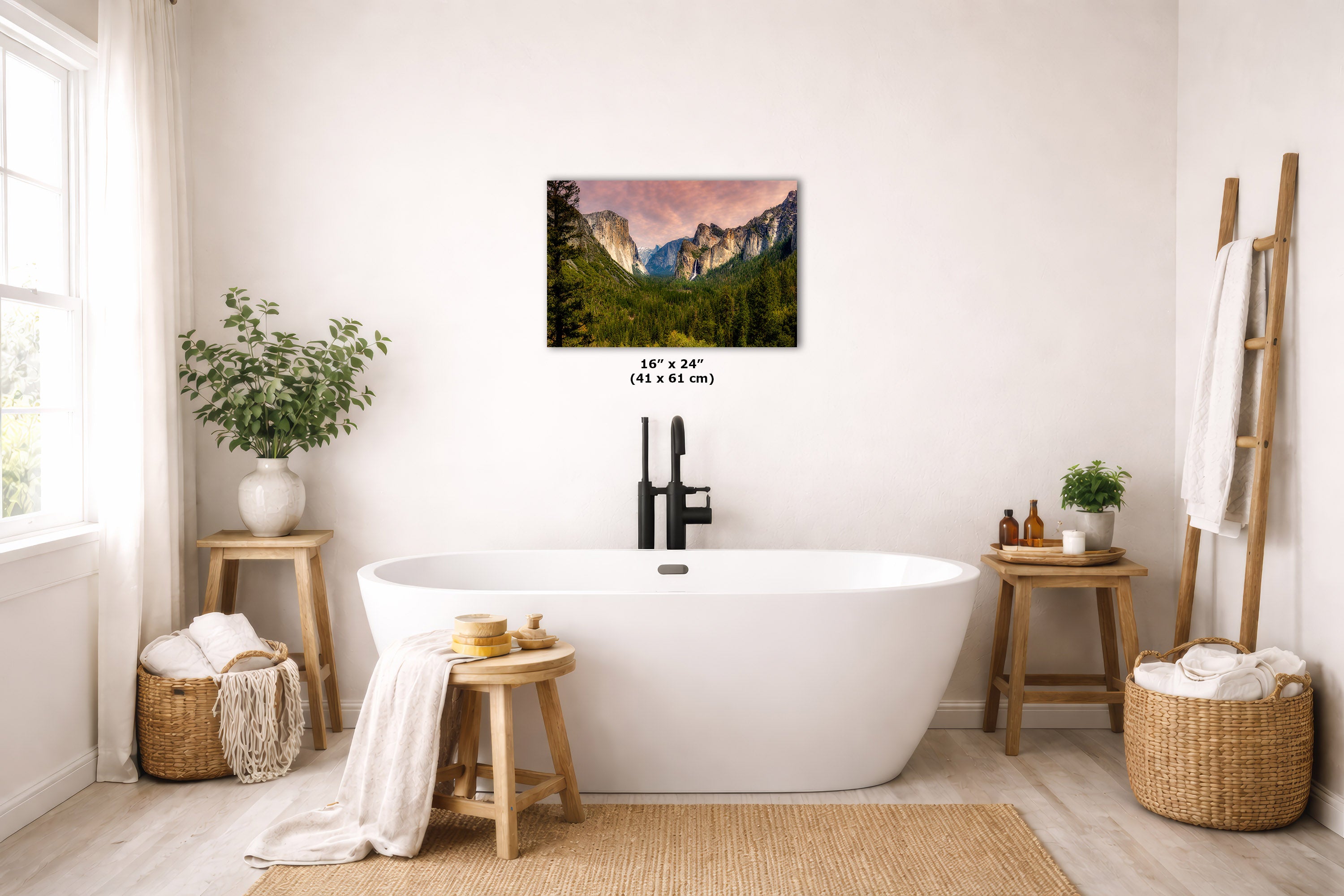 A wall art print depicting a mountain landscape with a pink and orange sunset sky, displayed above a white freestanding bathtub in a bright, modern bathroom with wooden stools, plants, and woven baskets.