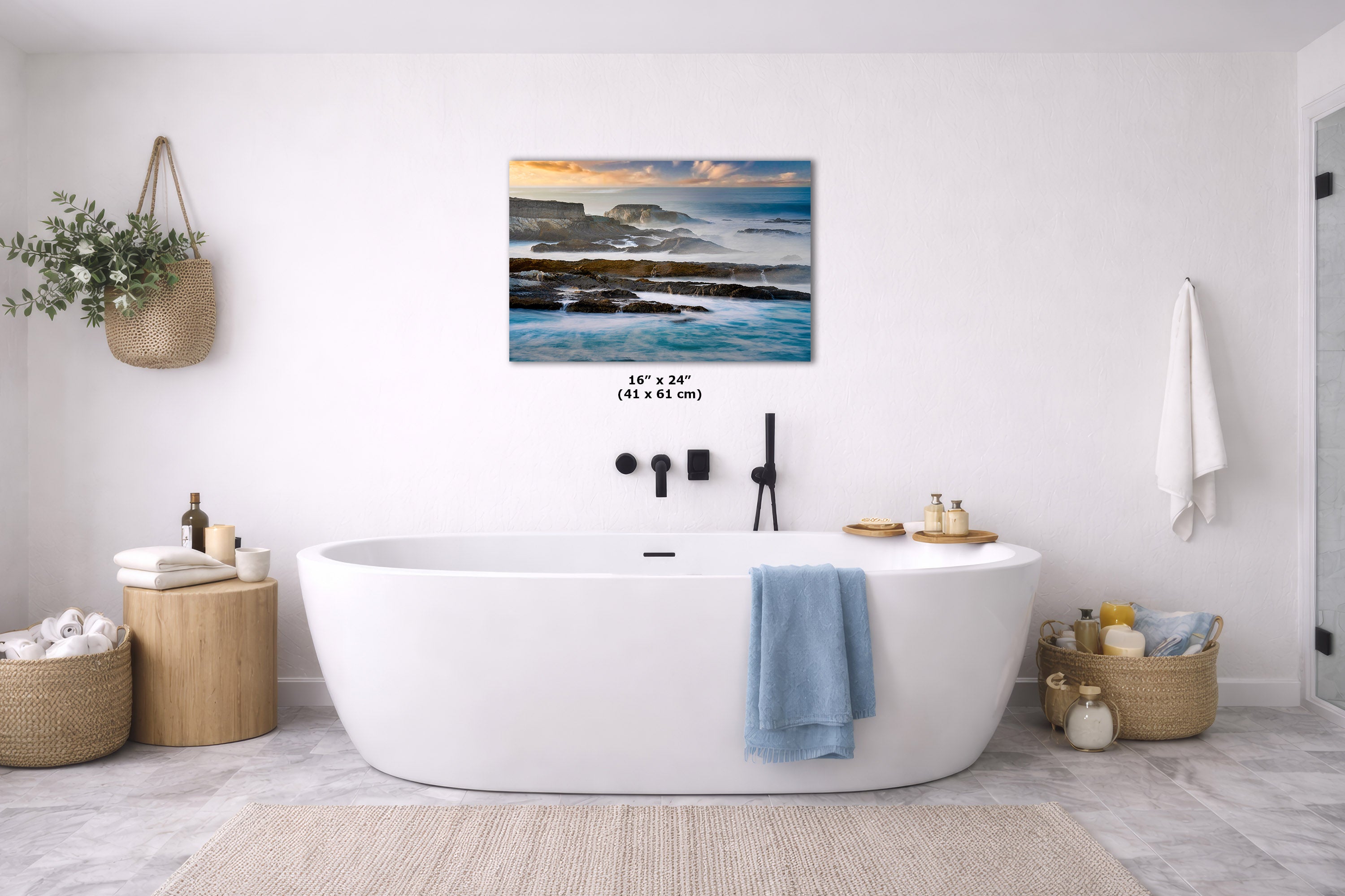 A wall art print depicting a coastal seascape with blue water, dark rocks, and a warm sunset sky, displayed above a white freestanding bathtub in a bright modern bathroom. Small size text reading 16 x 24" (41 x 61 in) is visible beneath the artwork.