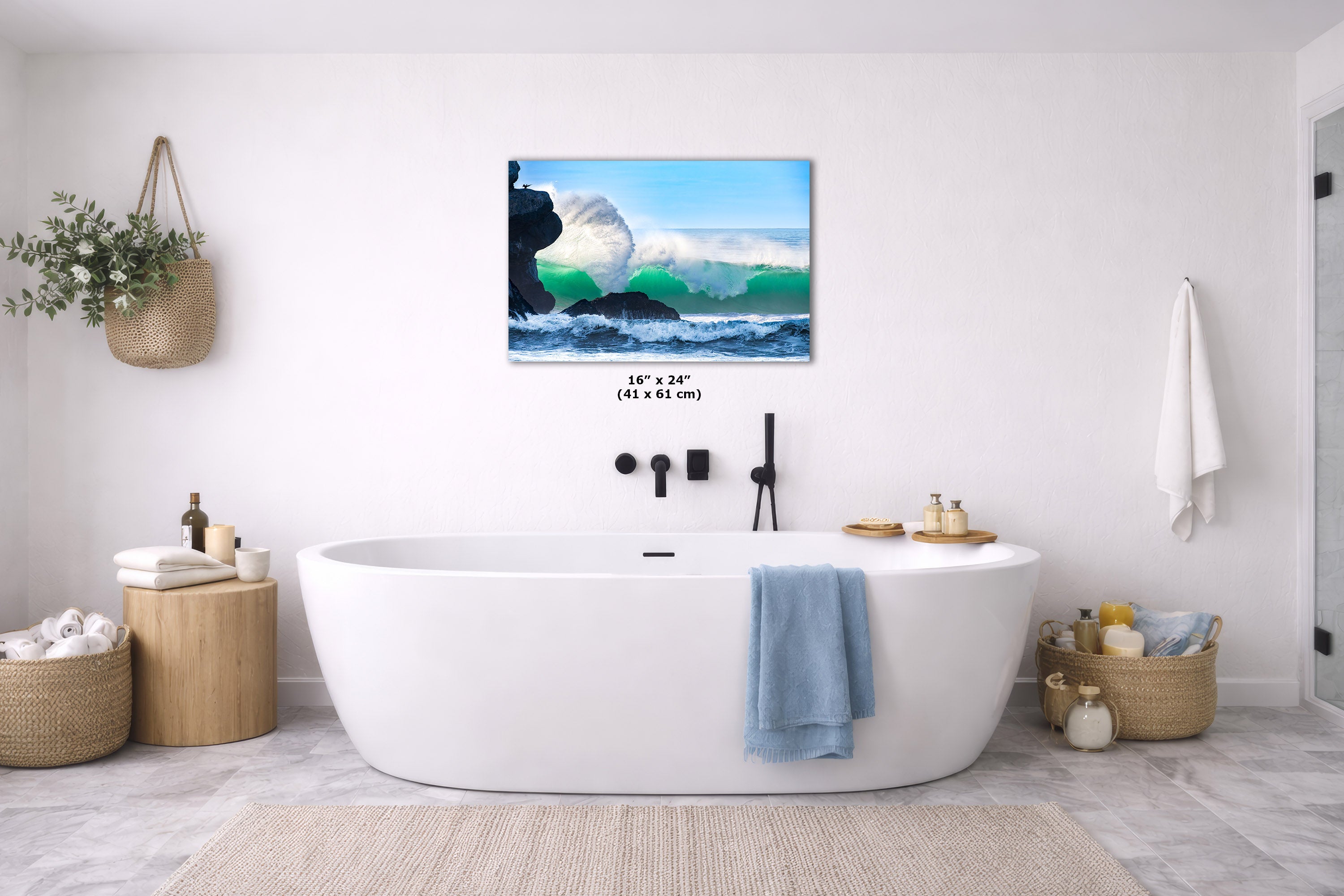 A wall art print depicting a dramatic ocean wave crashing over dark rocks under a blue sky, displayed above a white freestanding bathtub in a bright, modern bathroom. The print includes size text reading 16 x 24" (41 x 61 cm).