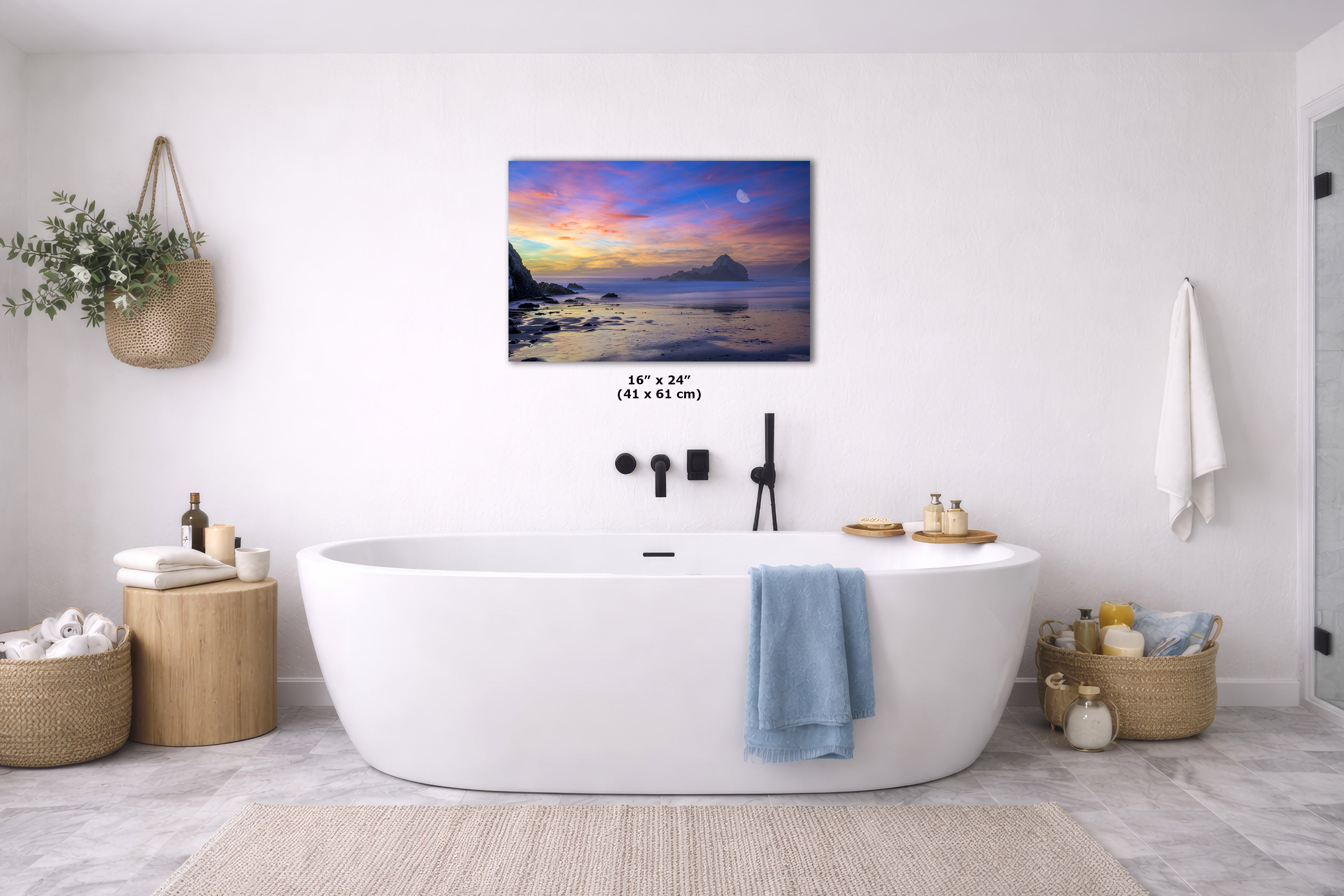 A wall art print showing a colorful sunset over water hangs above a white freestanding bathtub in a bright, modern bathroom. The print depicts a vivid sky with purple, pink, orange, and blue tones reflecting on the water and dark rocky shoreline.