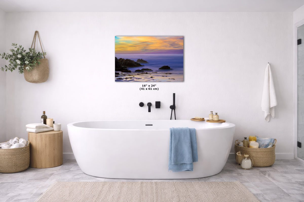 A rectangular wall art print showing a coastal sunset scene in yellow, orange, purple, and blue tones, displayed above a white freestanding bathtub in a bright modern bathroom. Small size text reading 16 x 24 in is printed near the artwork.