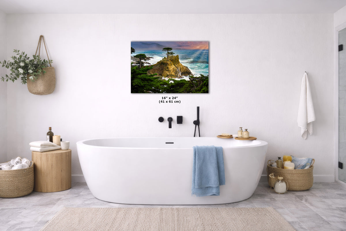 A coastal landscape wall art print hangs above a white freestanding bathtub in a bright, modern bathroom. The artwork shows a rocky shoreline with green trees, blue water, and a sunset sky.