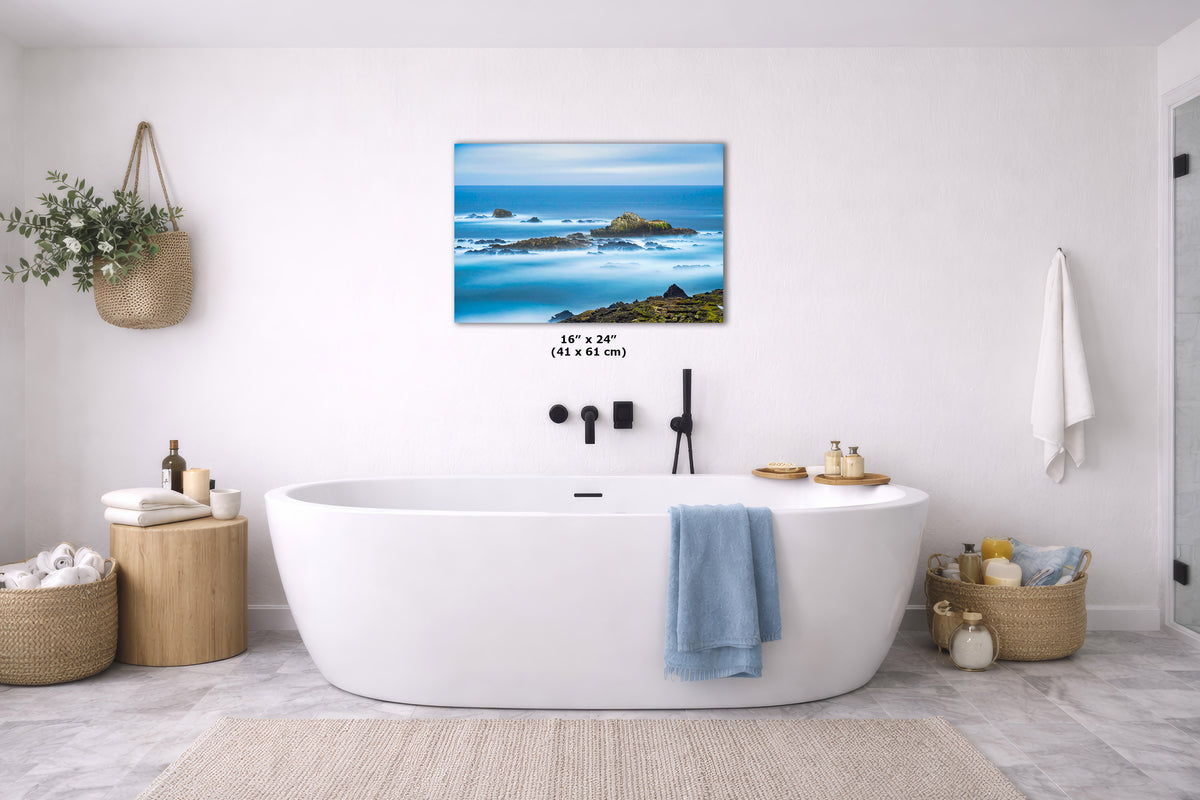 A modern bathroom scene with a freestanding white bathtub, blue towels, woven baskets, and a framed ocean landscape print centered on the wall. The print shows blue water, dark rocks, and a bright horizon, with size text beneath it reading 48 x 72 in.