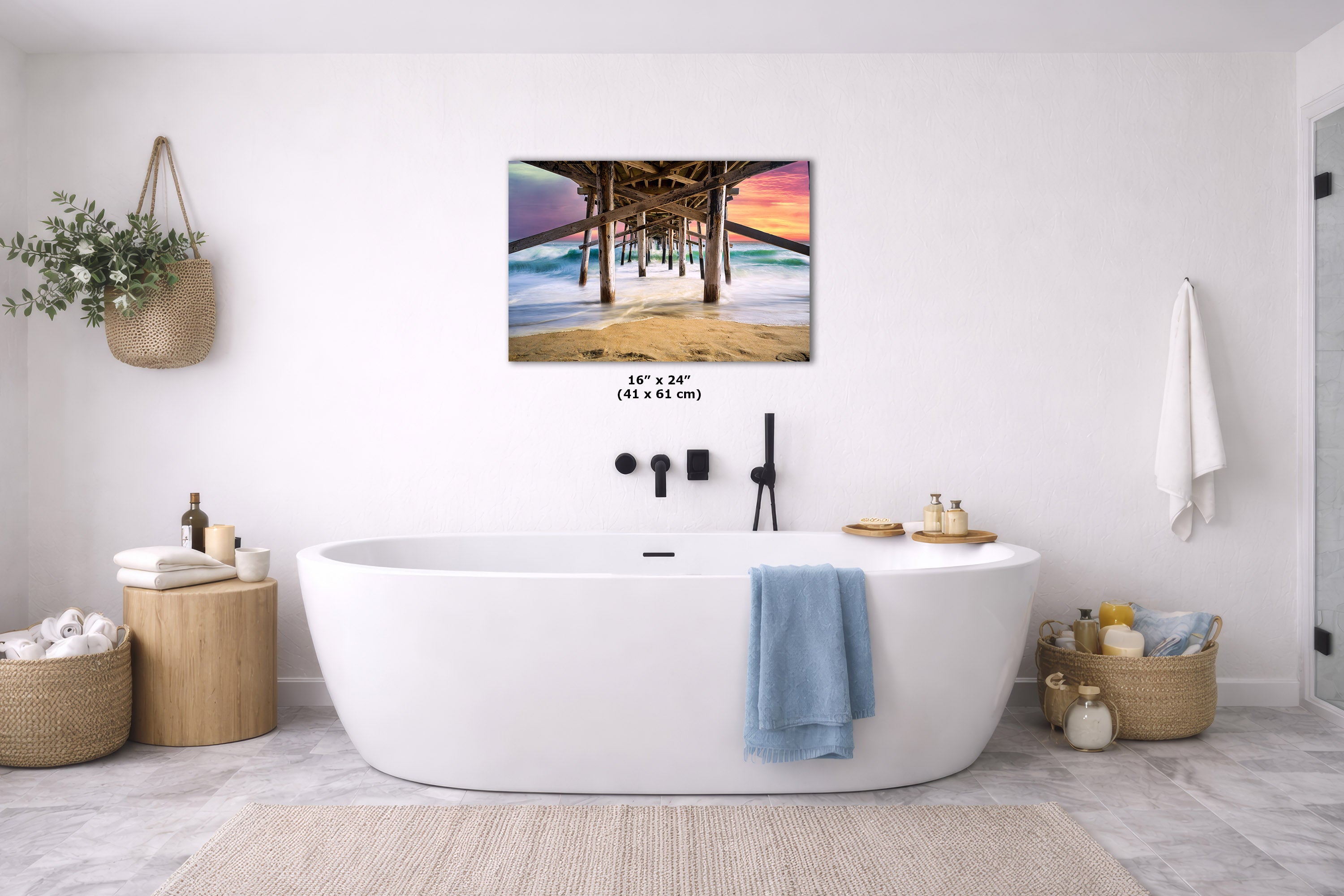 A wall art print showing a pier scene with colorful sunset sky, displayed above a white freestanding bathtub in a bright modern bathroom. The print includes size text reading 16 x 24" (41 x 61 cm).