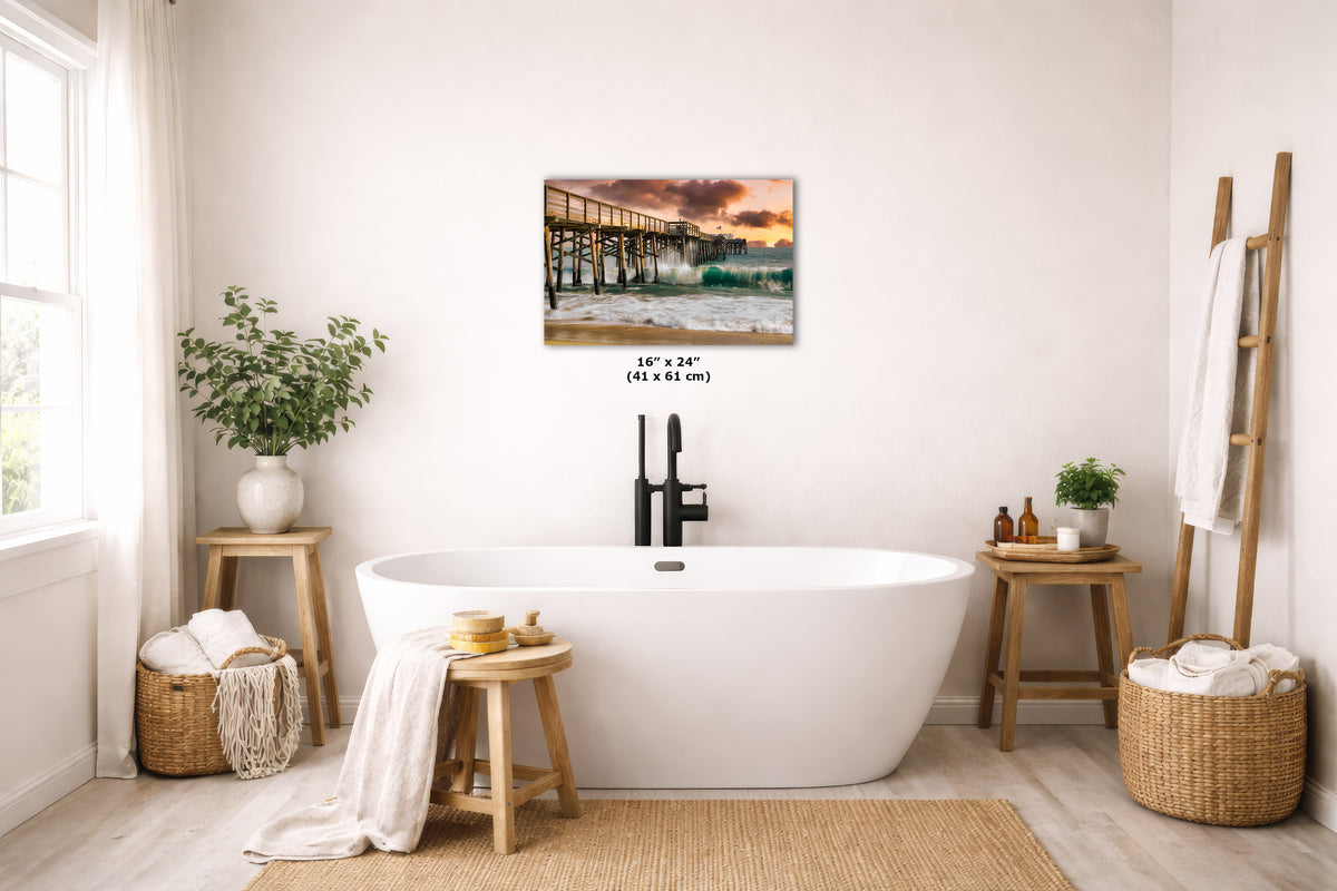 A wall art print depicting a sunset pier over ocean waves is displayed above a freestanding white bathtub in a bright, neutral-toned bathroom. The print includes size text reading 16 x 24 (41 x 61 cm).