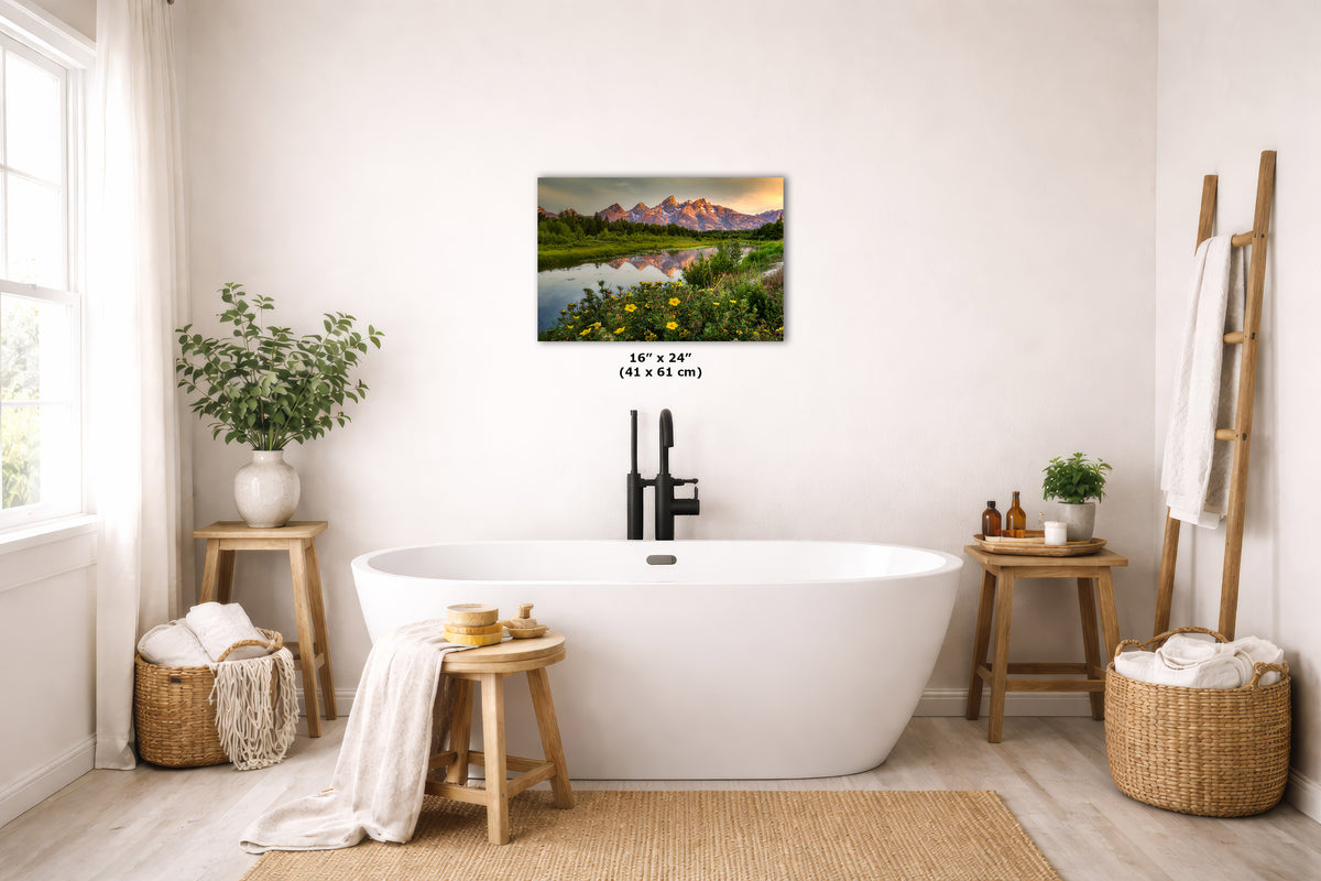 A freestanding white bathtub in a bright, neutral-toned bathroom with a forest wall art print above it. The scene includes wooden stools, woven baskets, plants, and a ladder-style towel rack.