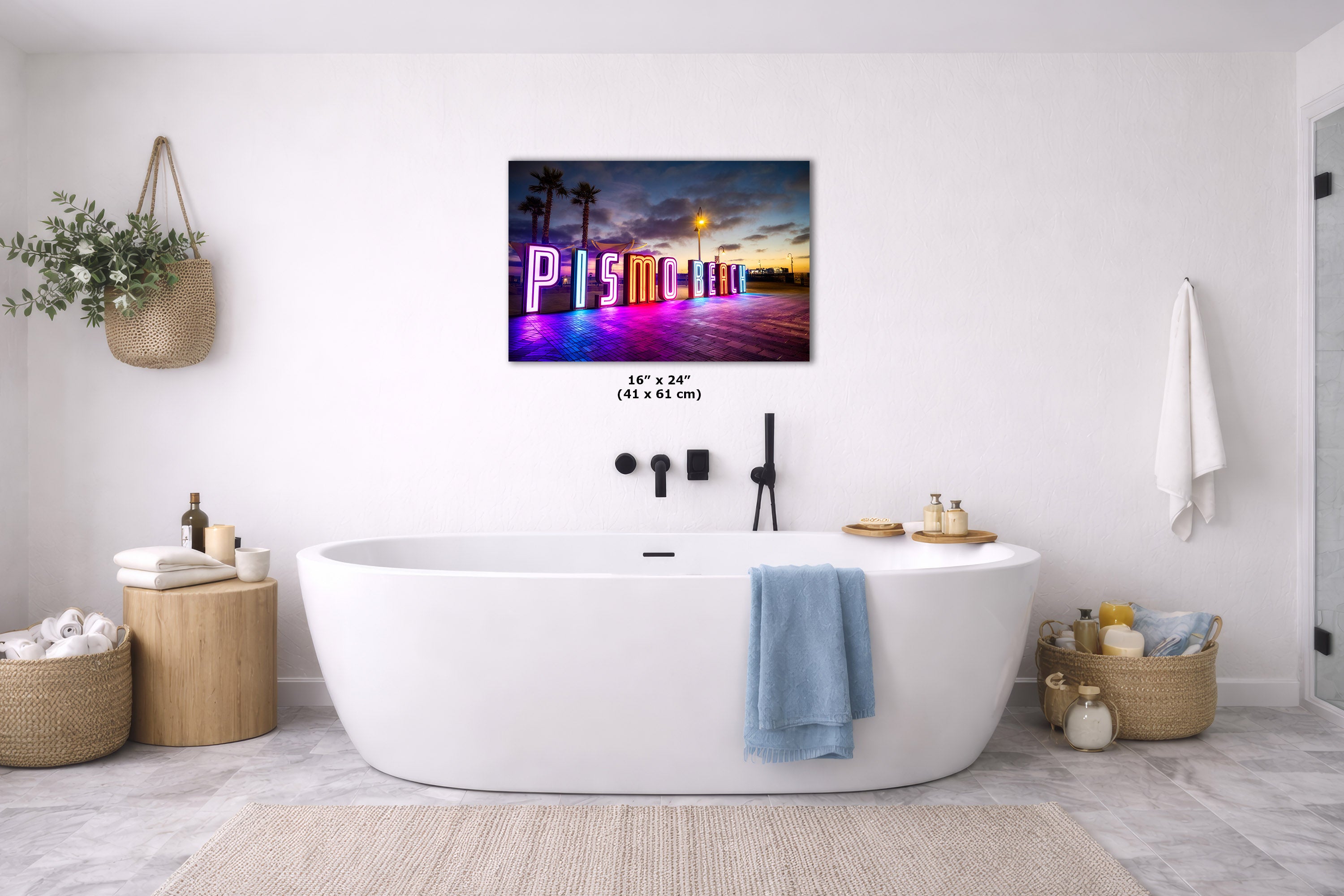 A freestanding oval bathtub in a bright white bathroom, with a colorful framed wall art piece above it showing a neon-style 'Pismo Beach' sign at sunset. The room includes a blue towel draped over the tub, woven baskets, and light wood side tables.