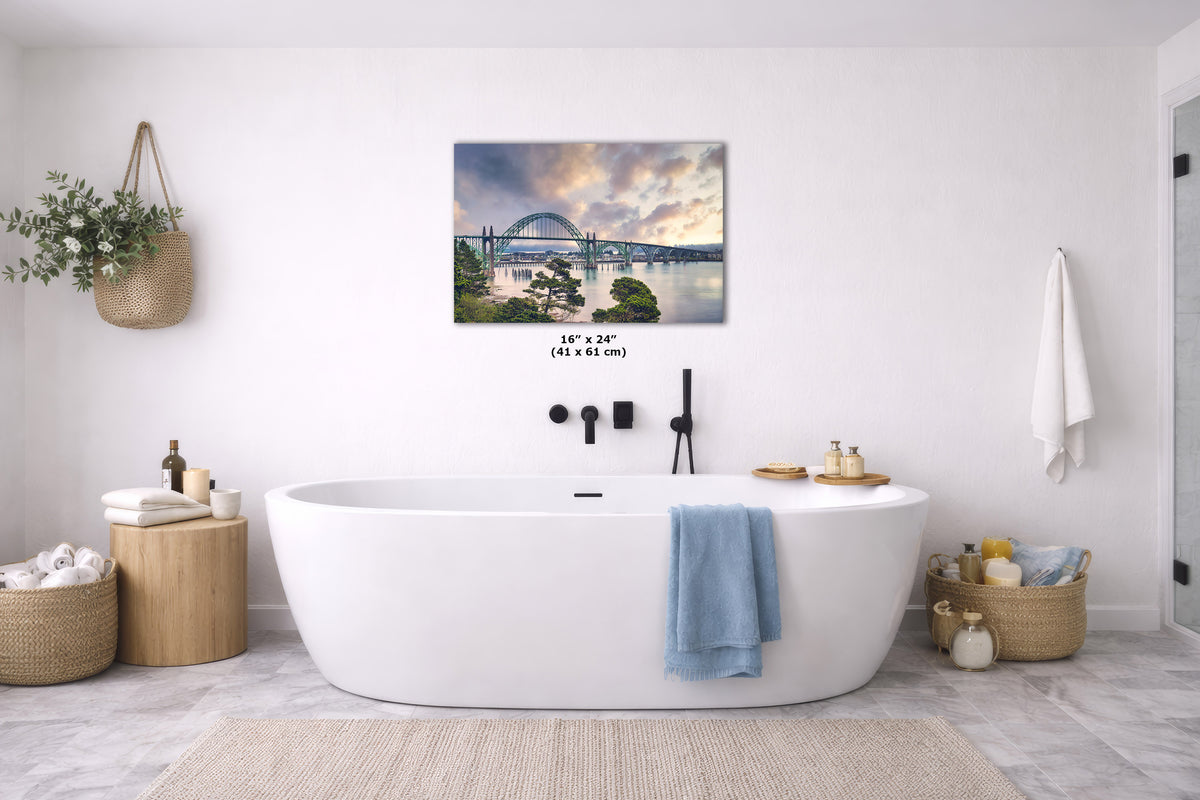 A freestanding oval bathtub in a bright white bathroom, with a framed landscape wall art print above it showing a bridge over water at sunset. The print includes size text reading 16 x 24 inches.