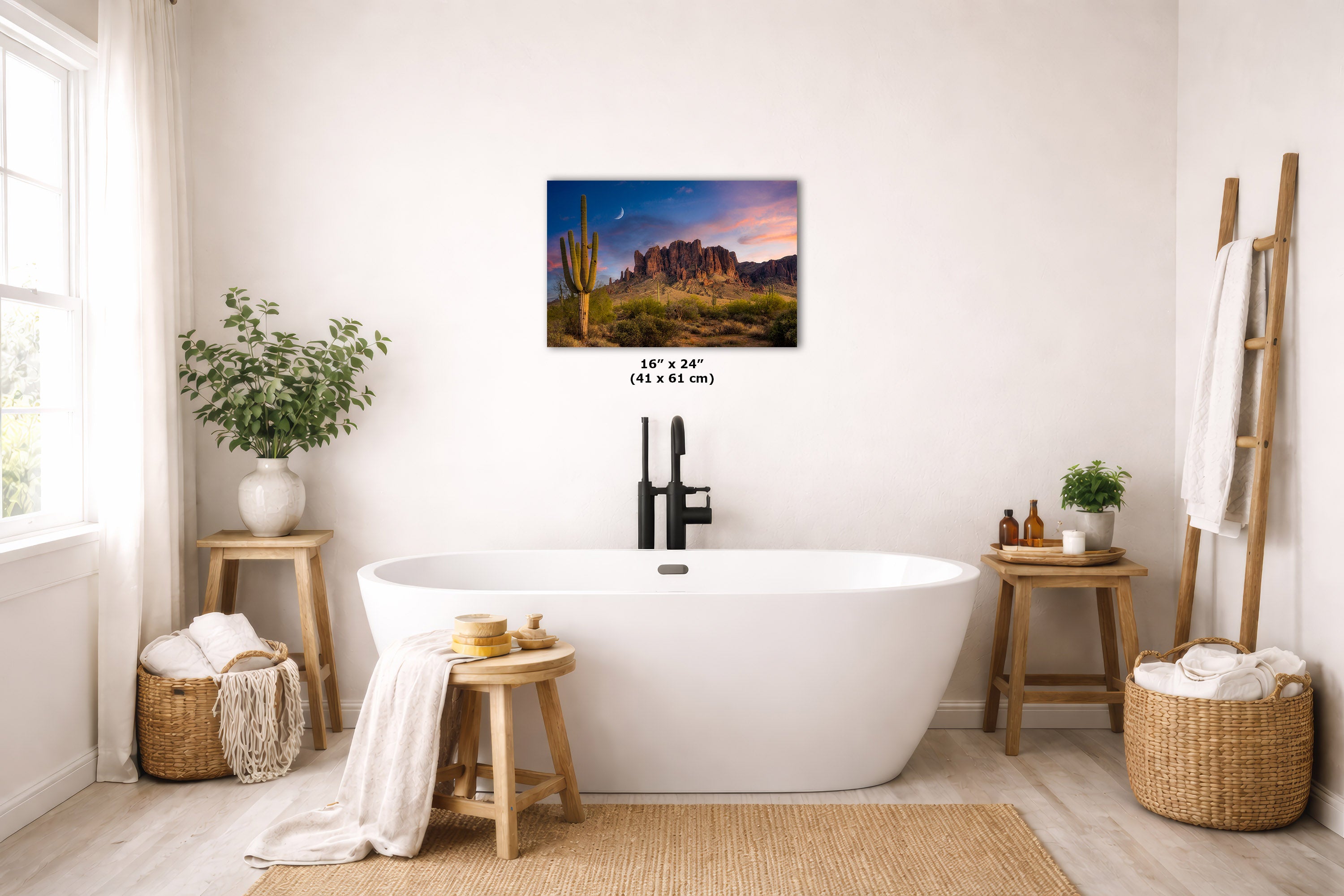 A desert landscape wall art print hangs above a white freestanding bathtub in a bright, neutral-toned bathroom. The artwork depicts red rock desert mountains at sunset with cacti.