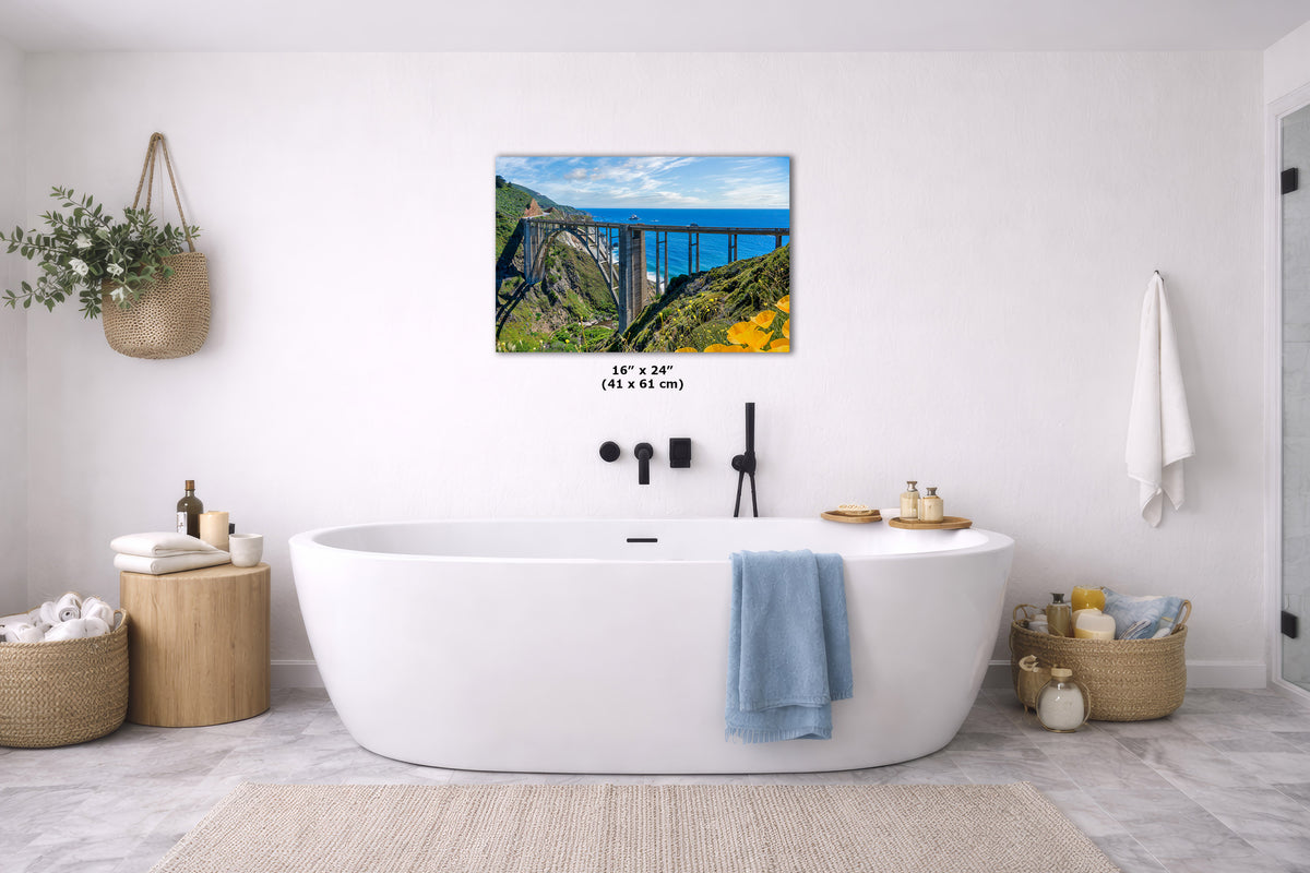 A wall art print depicting a coastal bridge over green cliffs beneath a blue sky, displayed above a white freestanding bathtub in a bright, modern bathroom. The print includes size text reading 16 x 24 inches.