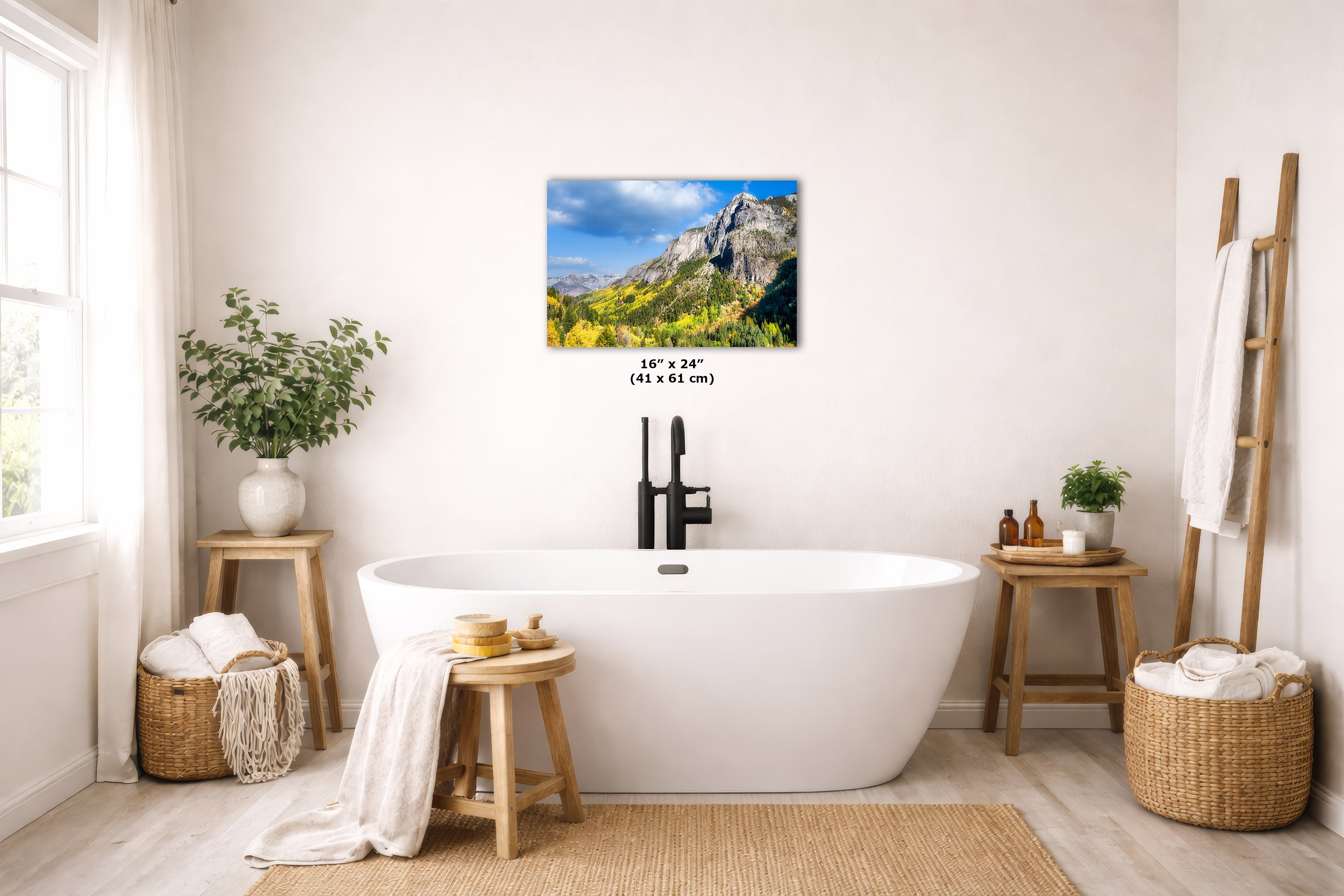 A wall art print showing a mountain landscape with blue sky and green-yellow foliage, displayed above a white freestanding bathtub in a bright, neutral-toned bathroom. Small text beneath the print reads 16 x 61 (41 x 61 cm).