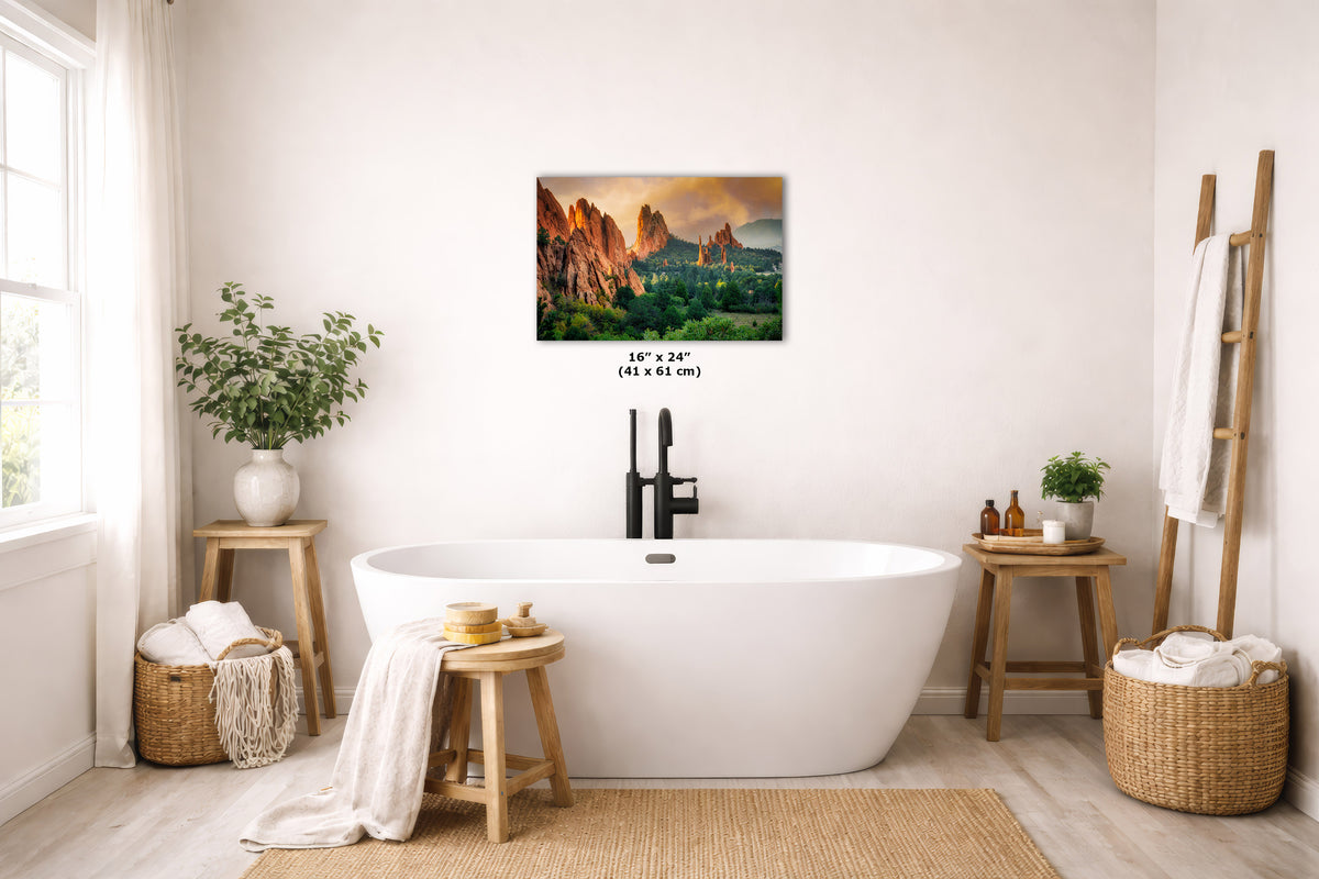 A wall art print depicting a mountain landscape with a pink and orange sunset sky, displayed above a white freestanding bathtub in a bright, modern bathroom with wooden stools, plants, and woven baskets.