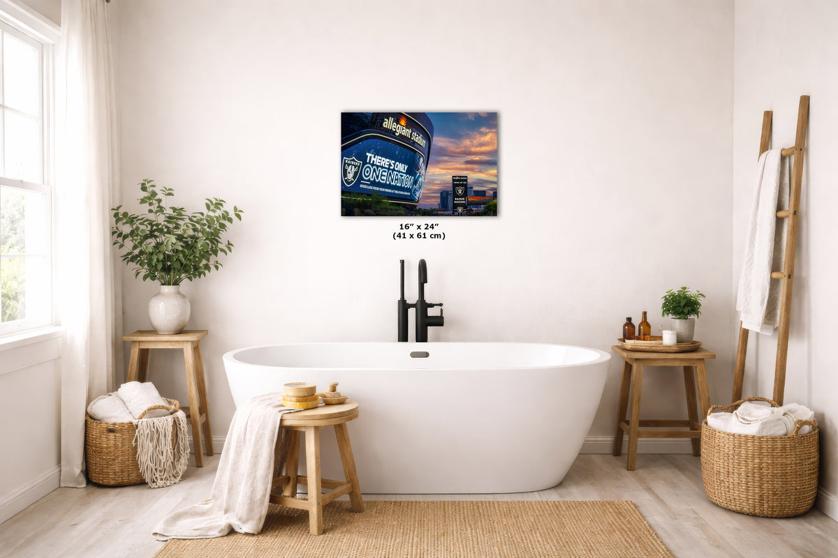A wall art print hangs above a white freestanding bathtub in a bright, neutral-toned bathroom. The artwork depicts the Las Vegas Raiders Allegiant stadium at sunset. 