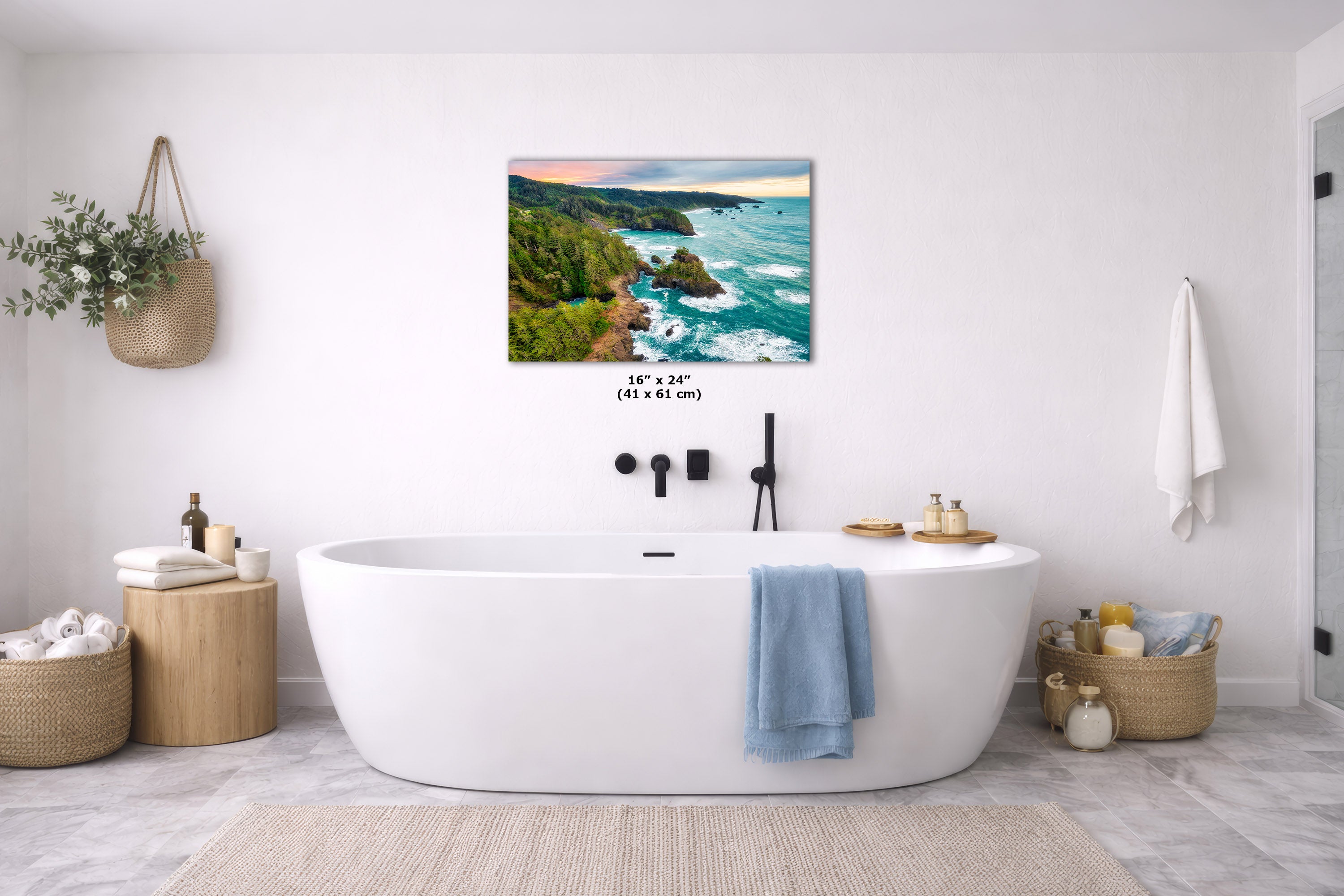 A coastal landscape wall art print hangs above a white freestanding bathtub in a bright, modern bathroom. The artwork shows a rocky shoreline with turquoise water and a sunset sky.