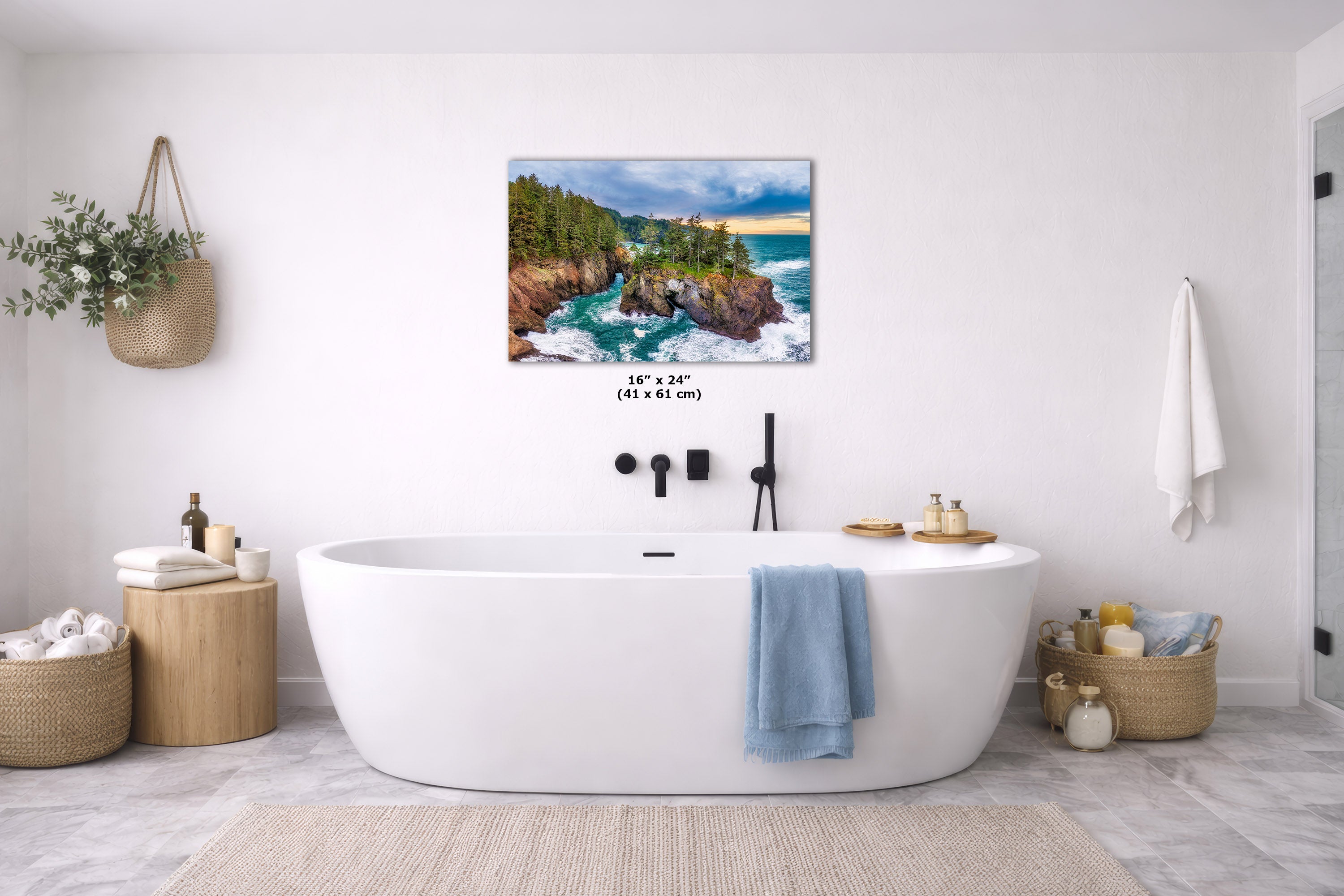 A coastal landscape wall art print hangs above a white freestanding bathtub in a bright, modern bathroom. The artwork depicts rocky cliffs, turquoise water, and a blue sky.