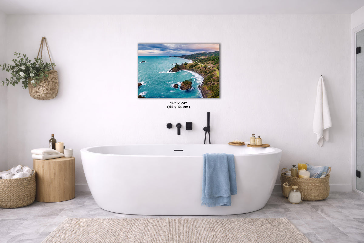 A coastal landscape wall art print hangs above a white freestanding bathtub in a bright, modern bathroom. The artwork shows turquoise water, rocky shoreline, and green hills under a blue sky.