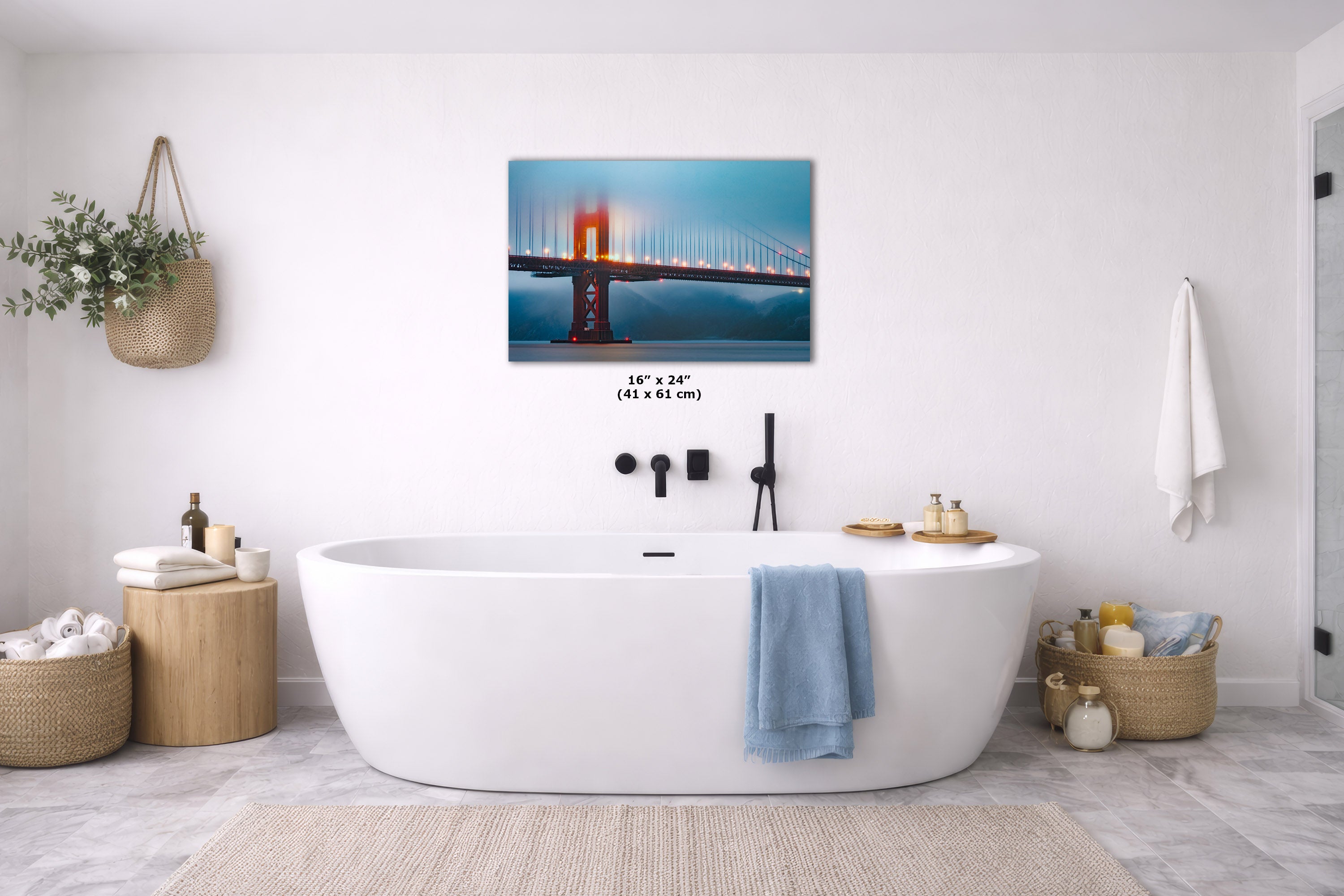 A wall art print of the Golden Gate Bridge hanging above a white freestanding bathtub in a bright, modern bathroom. The print shows the bridge at dusk with blue water and orange-red light.