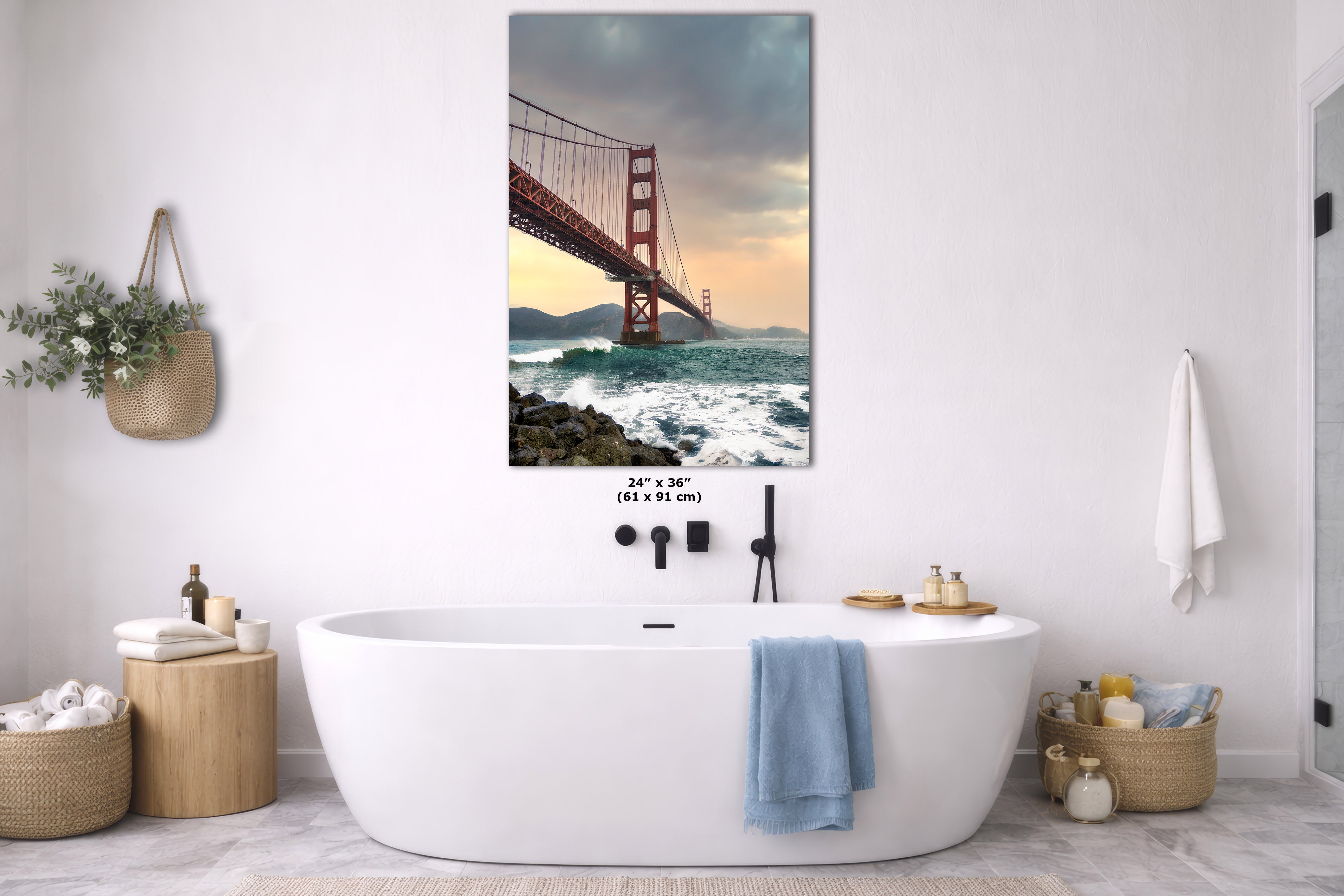 A framed wall art print of the Golden Gate Bridge over the ocean hangs above a white freestanding bathtub in a bright, modern bathroom. The print shows the red-orange bridge against a dramatic sky with waves below.