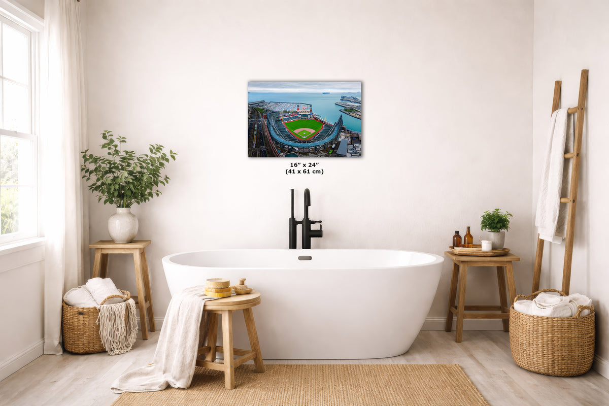 A wall art print showing an aerial view of the Oracle Park baseball stadium is displayed above a freestanding white bathtub in a bright, modern bathroom. The print includes size text below it reading 16 x 24 inches (41 x 61 cm).