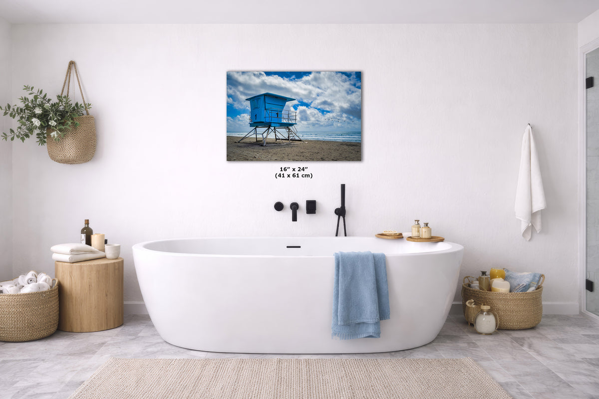 A wall art print showing a blue beach lifeguard tower under a cloudy sky, displayed above a white freestanding bathtub in a bright modern bathroom. The print includes size text reading 16 x 24 inches.