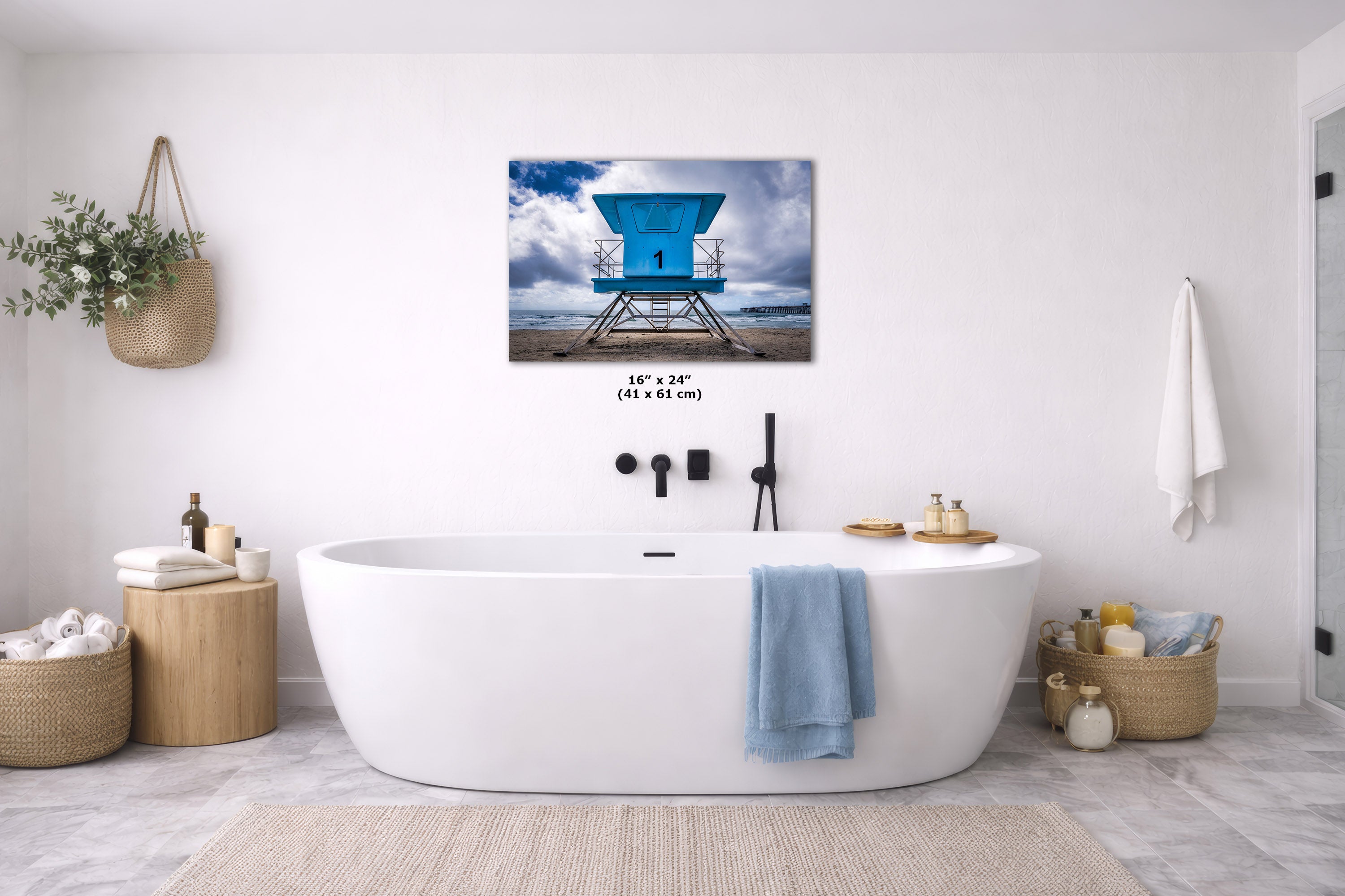A wall art print showing a bright blue lifeguard tower over water, displayed above a white freestanding bathtub in a bright, modern bathroom. The print includes size text reading 16" x 24" (41 x 61 in).